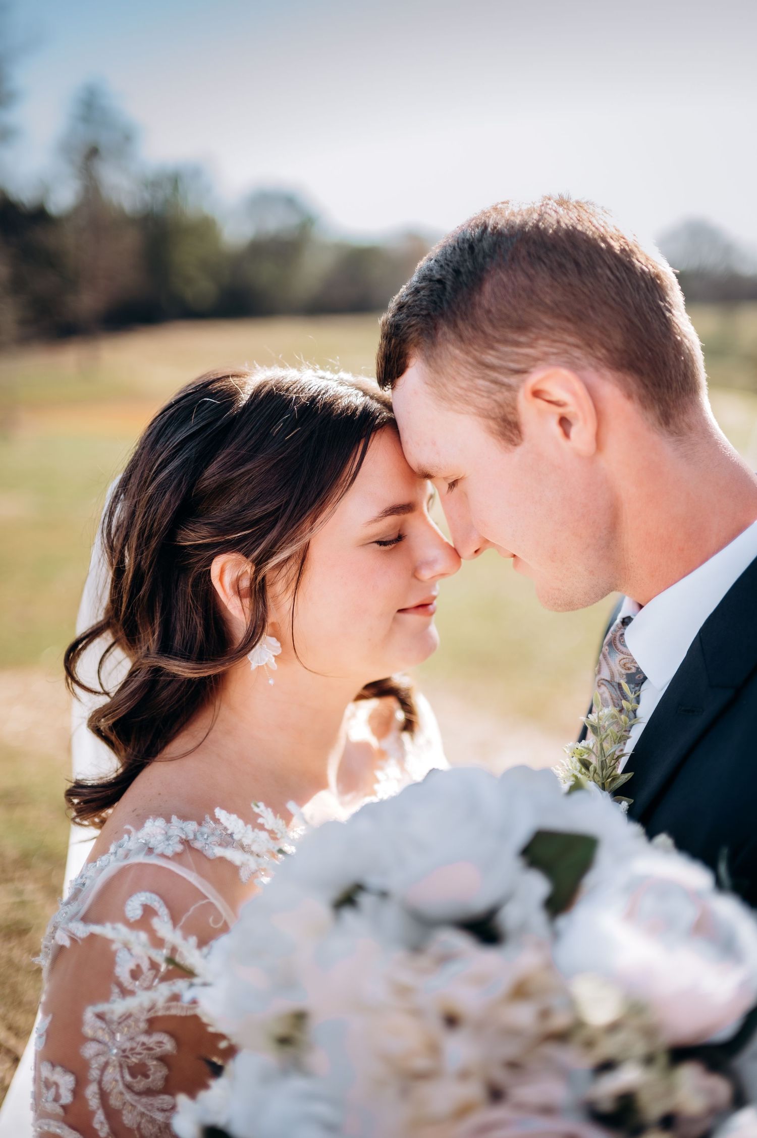 A bride and groom share a romantic forehead touch, bride holding white peony bouquet, wearing lace gown outdoors.