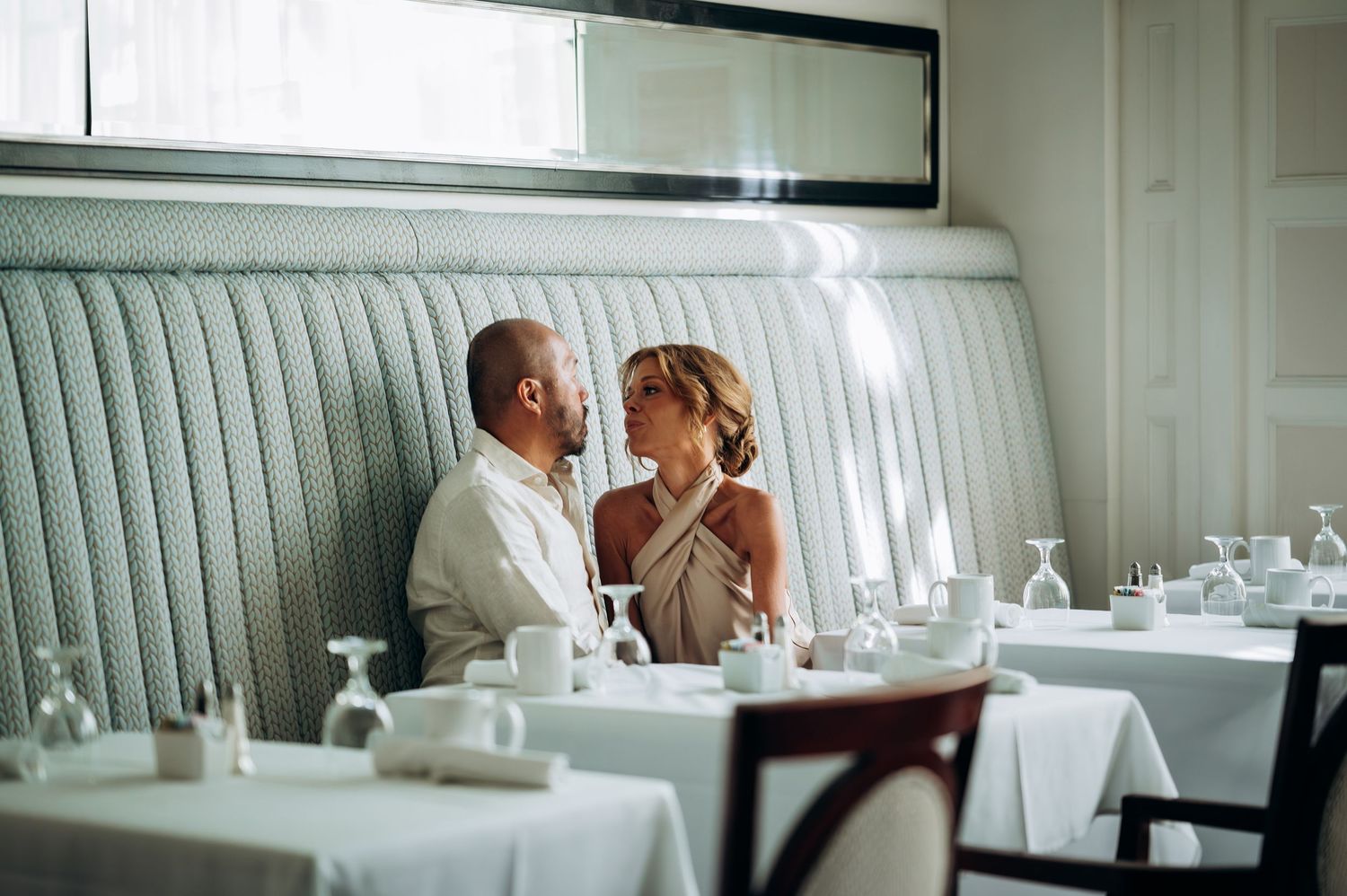 A romantic couple shares an intimate moment in a elegant restaurant booth, dressed in wedding attire.