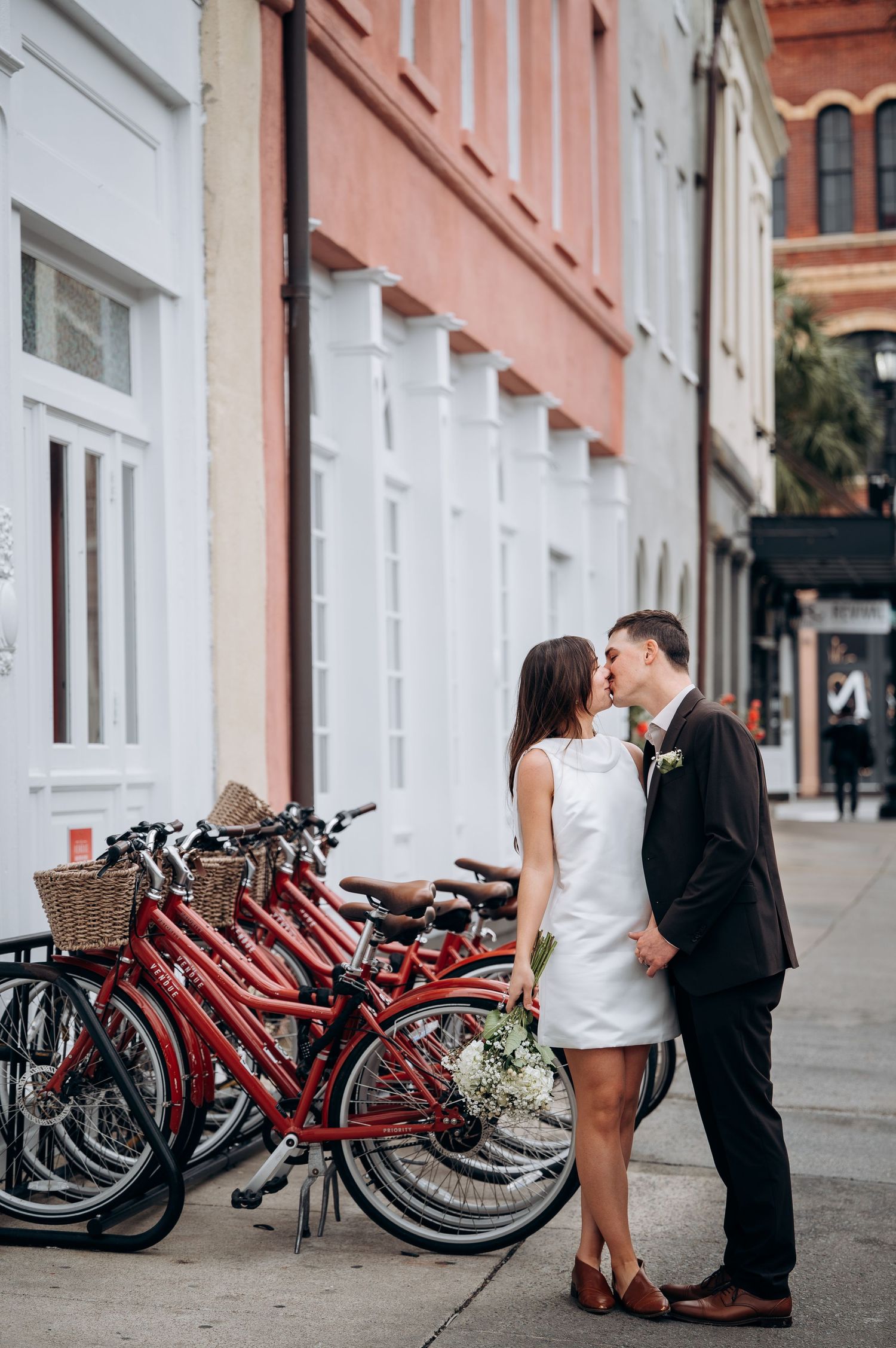 Wedding couple shares a kiss beside red bicycles on a charming city sidewalk.