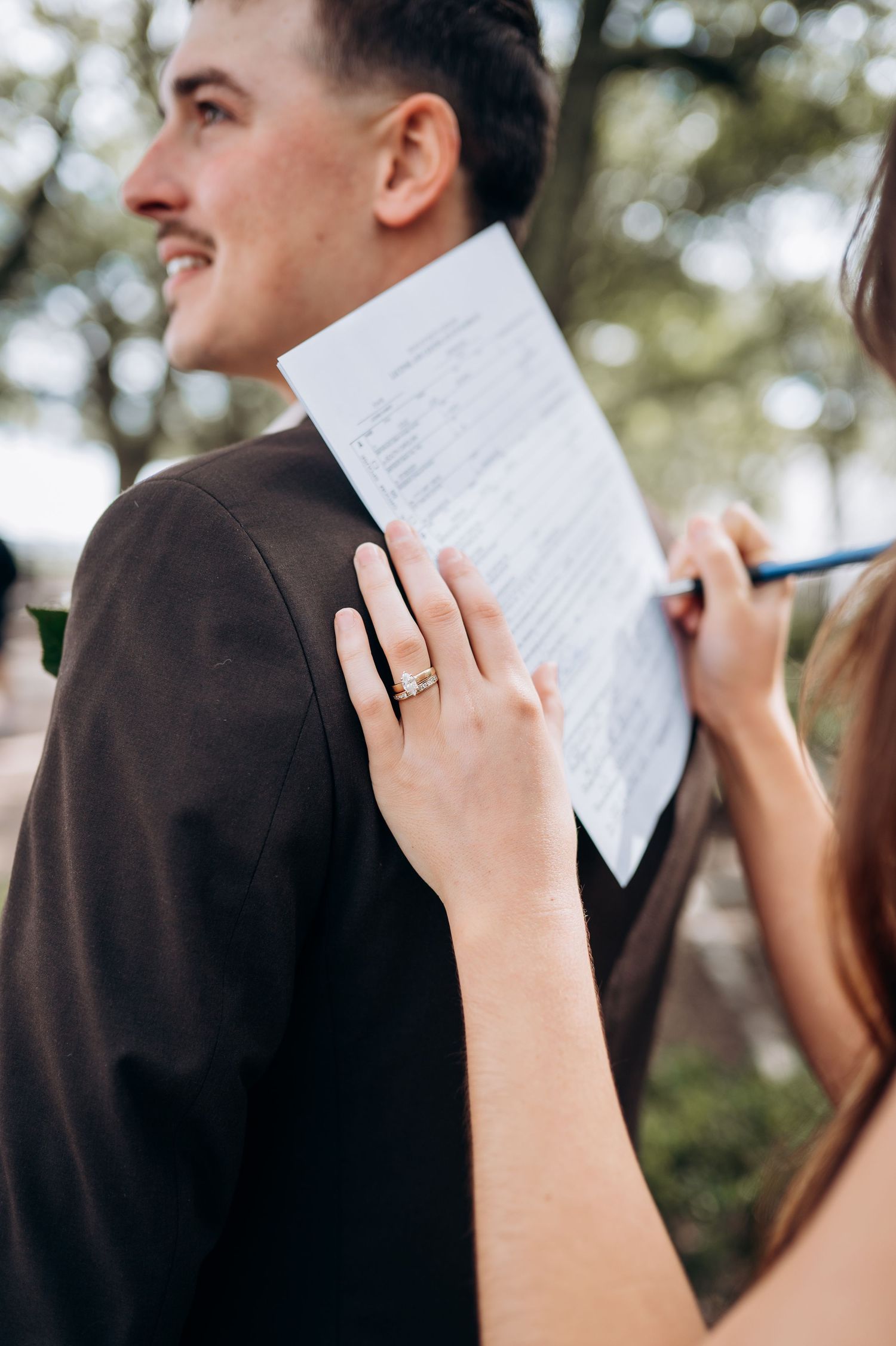 A smiling groom in a brown jacket holds wedding documents while a hand with a ring signs them outdoors.