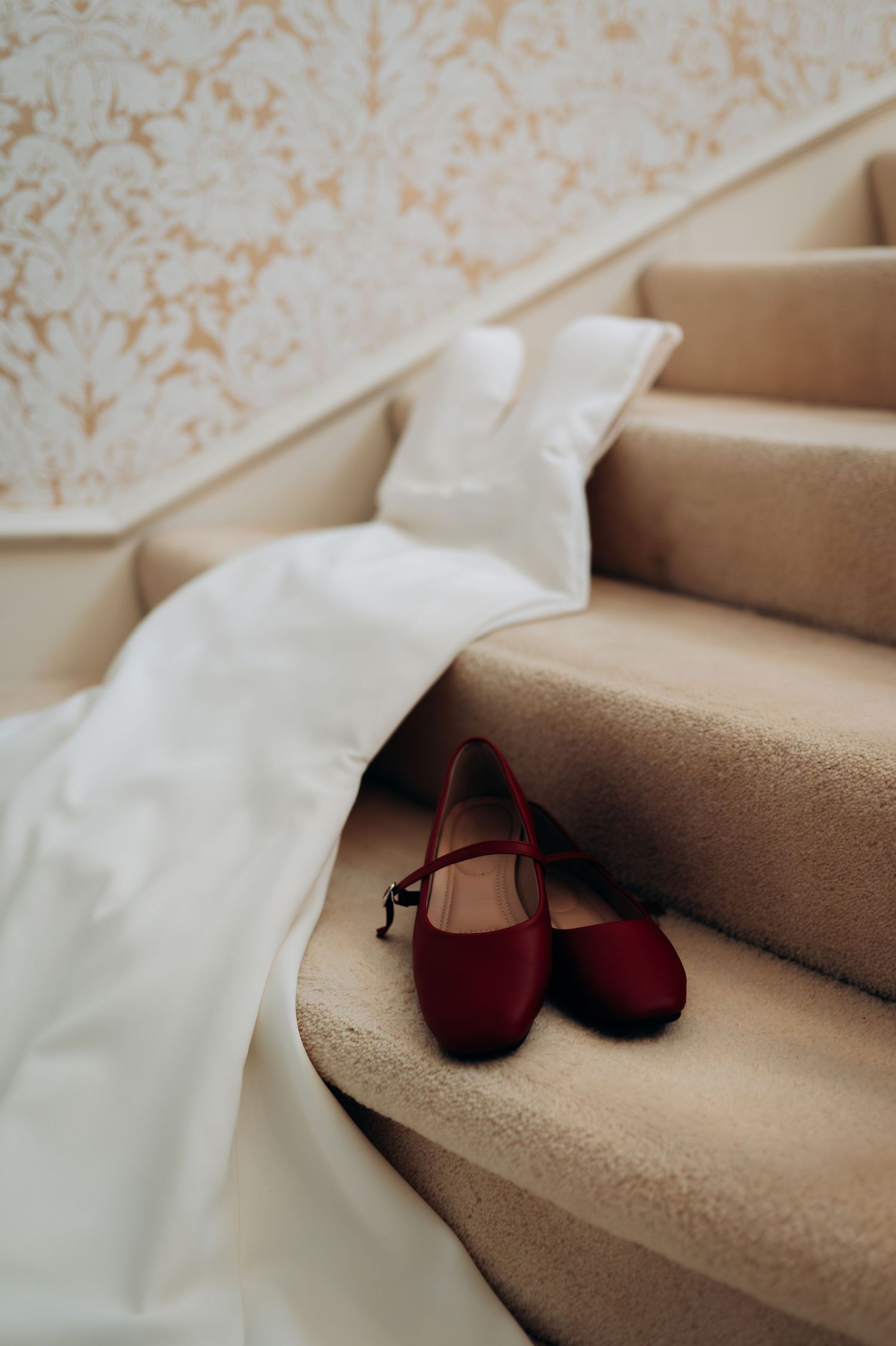 Red bridal ballet flats on carpeted stairs beside a flowing white wedding dress train.