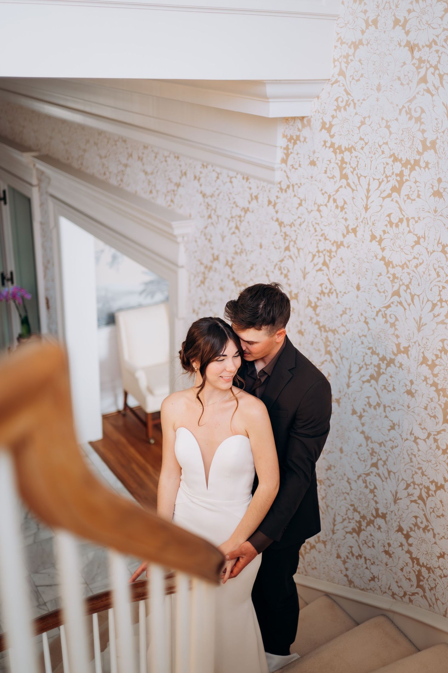 Groom embracing bride from behind on staircase with gold floral wallpaper backdrop.