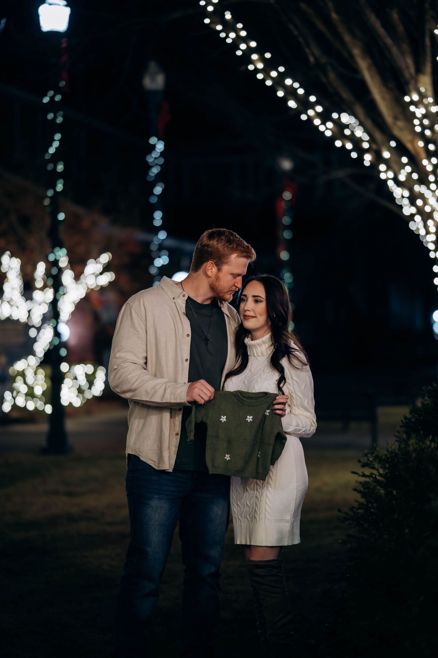 Couple holding tiny green baby outfit under twinkling holiday lights at night.