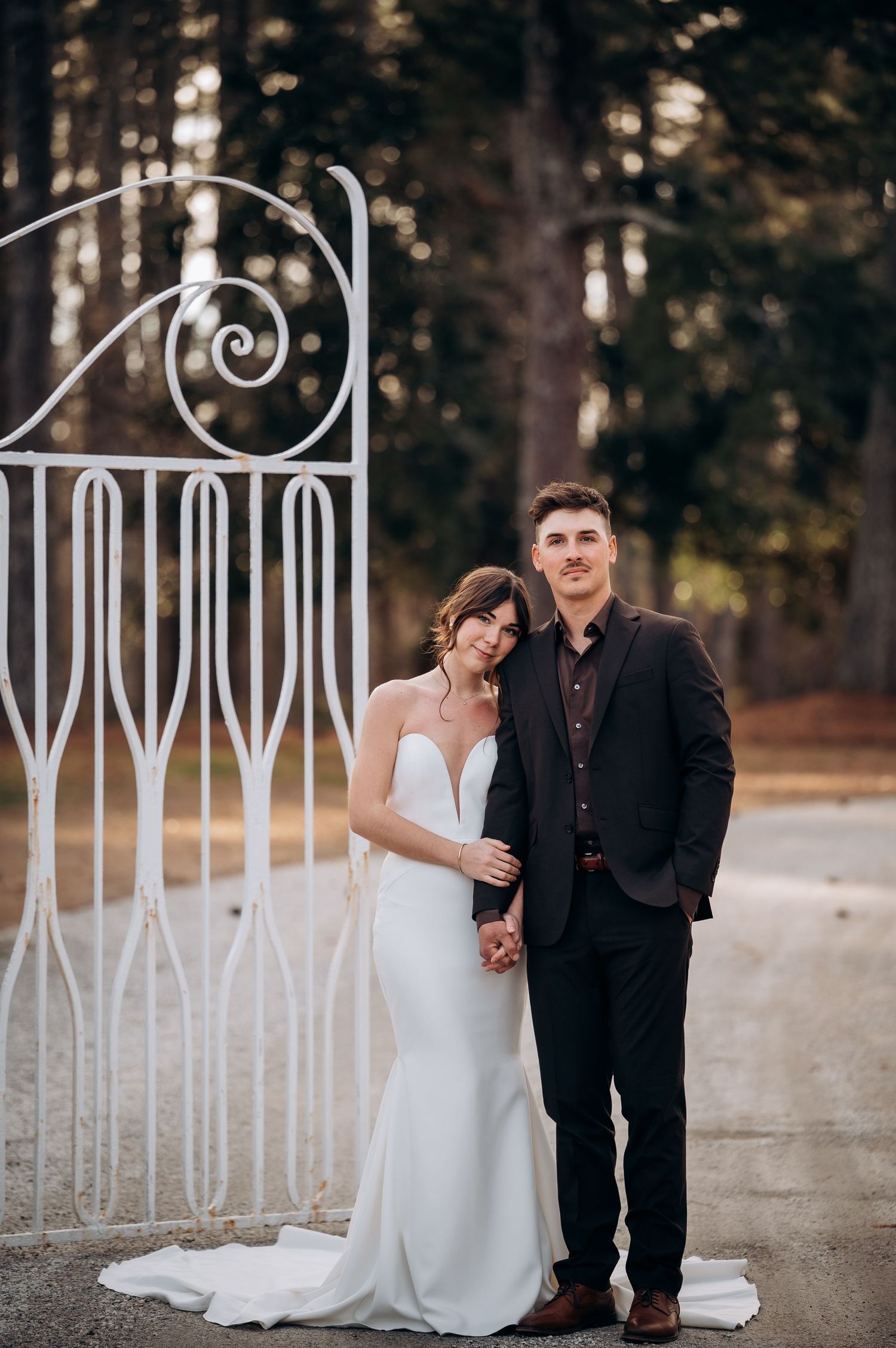 A couple in wedding attire poses against an ornate white metal fence in a wooded outdoor setting.