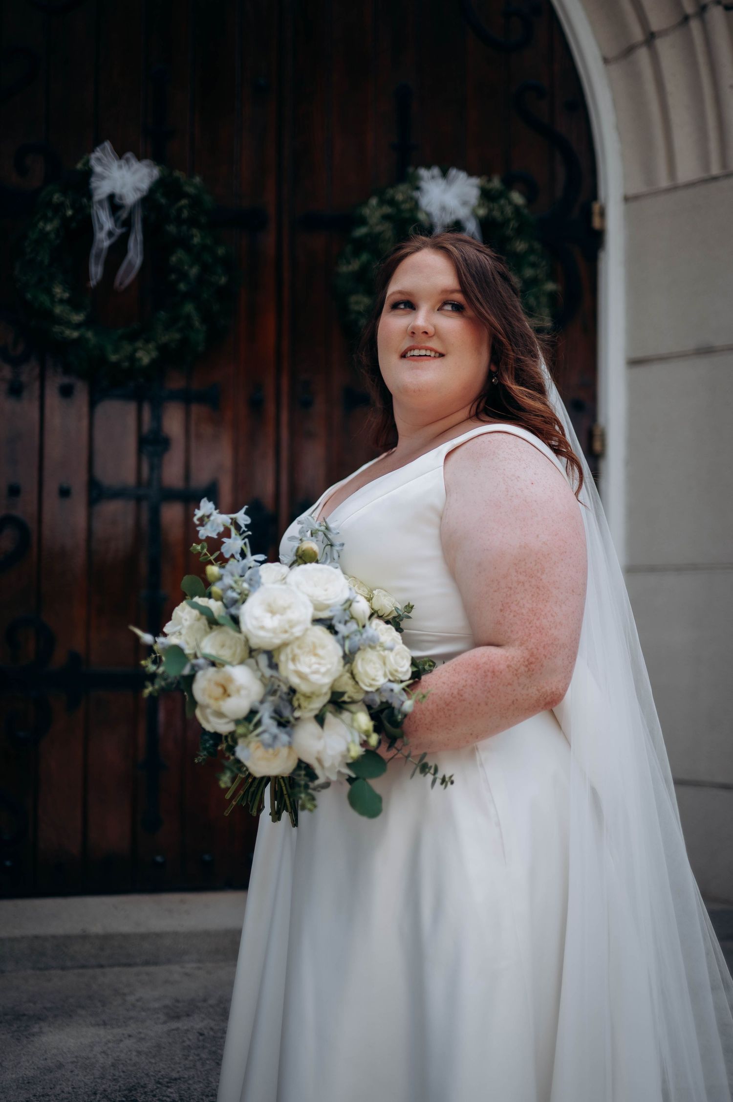 Bride in elegant white dress holds classic white and green bouquet against church backdrop.