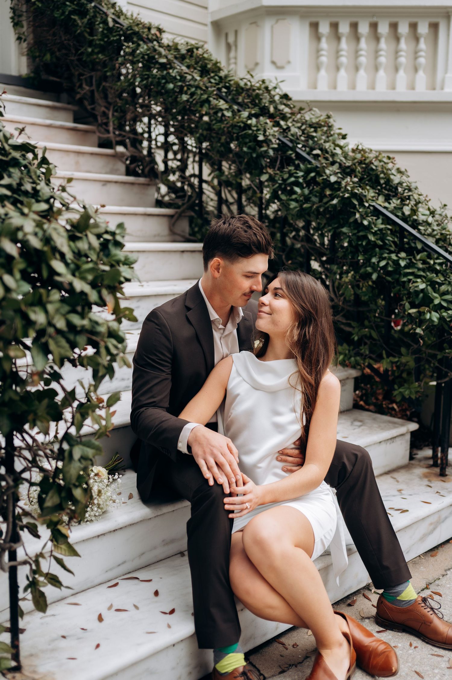 A couple shares intimate moments while sitting on white stone steps surrounded by lush greenery.