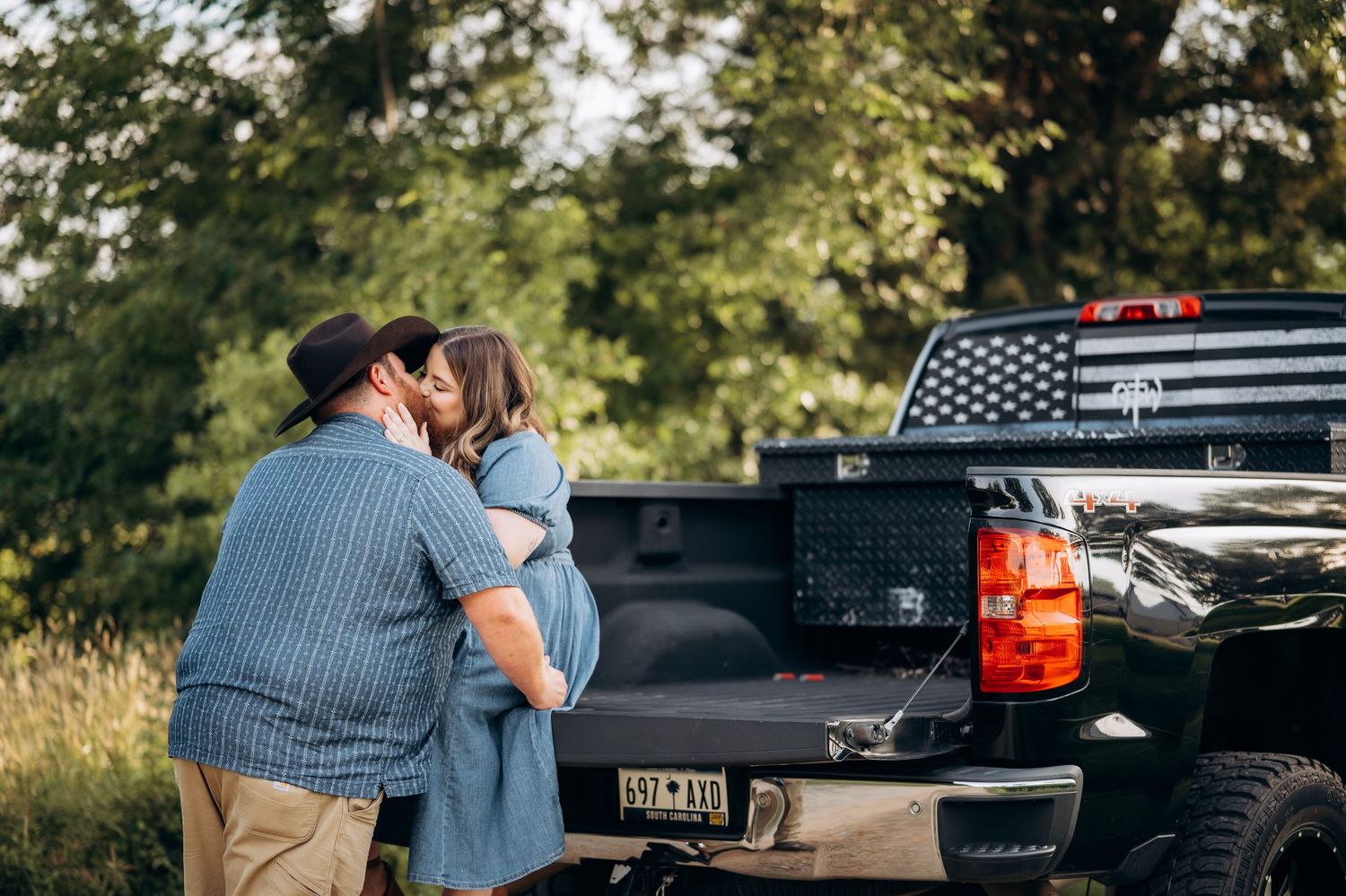 Couple sharing a romantic kiss beside a black pickup truck with an American flag decal in a lush outdoor setting.