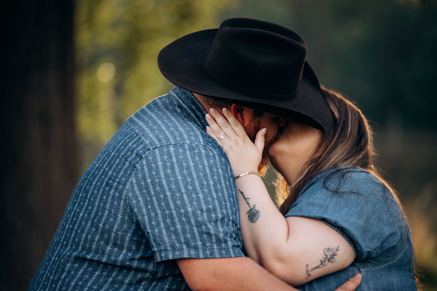 Couple sharing a romantic kiss outdoors, woman with tattoos and man wearing a black cowboy hat embracing.