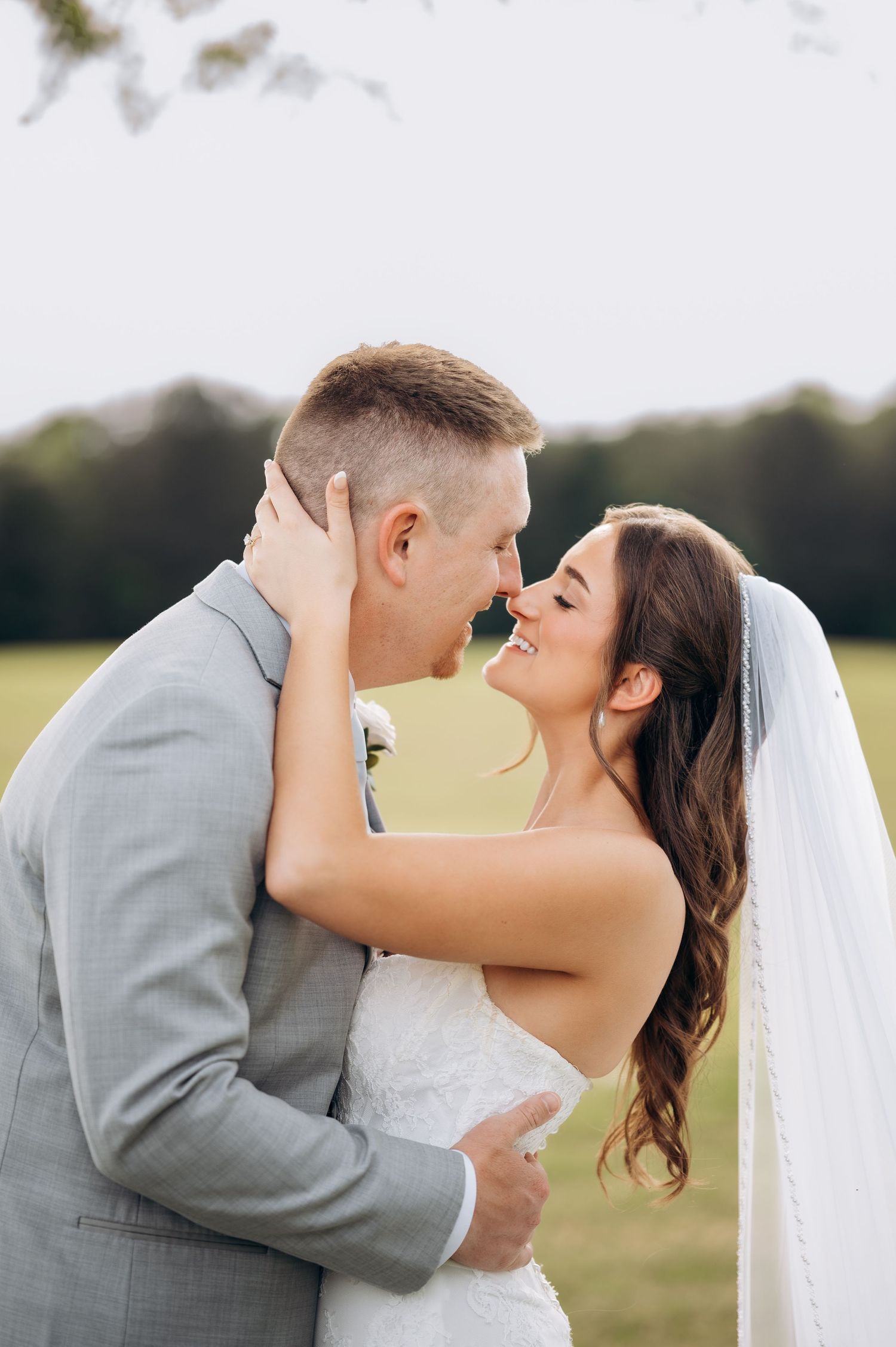A bride and groom share a romantic almost-kiss outdoors, she in a white gown with veil, he in a gray suit.