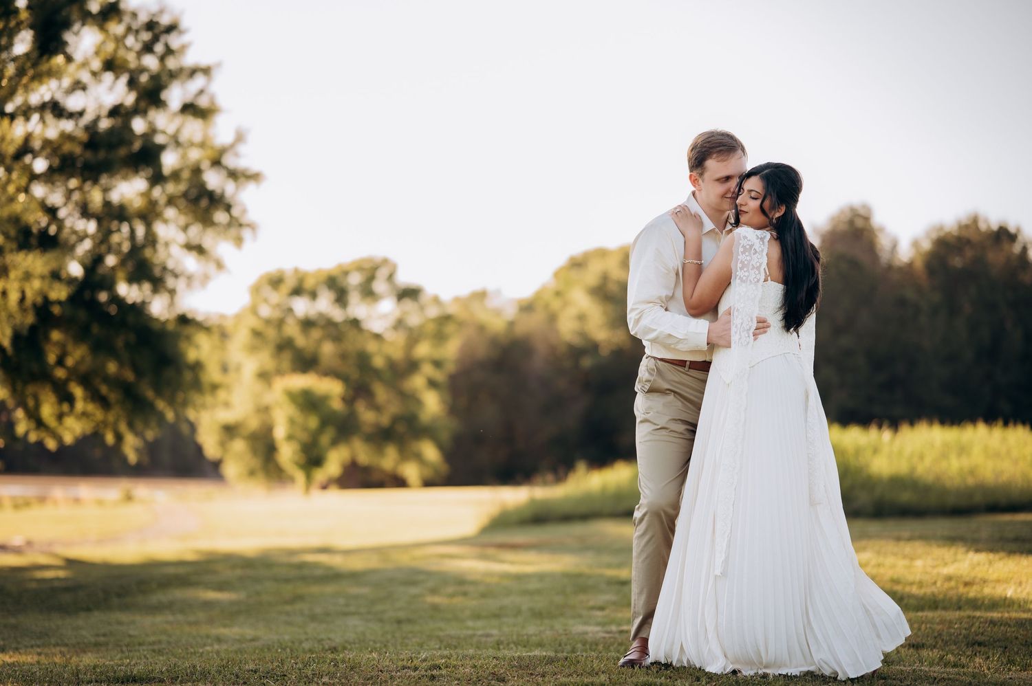 Couple embracing in wedding attire on lush green lawn at golden hour, bride in white gown, groom in tan suit.