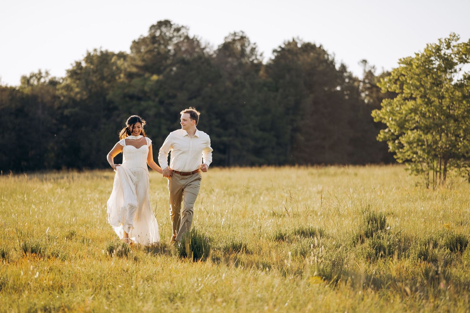 Bride and groom holding hands running through a sunlit meadow with lush green trees in the background.