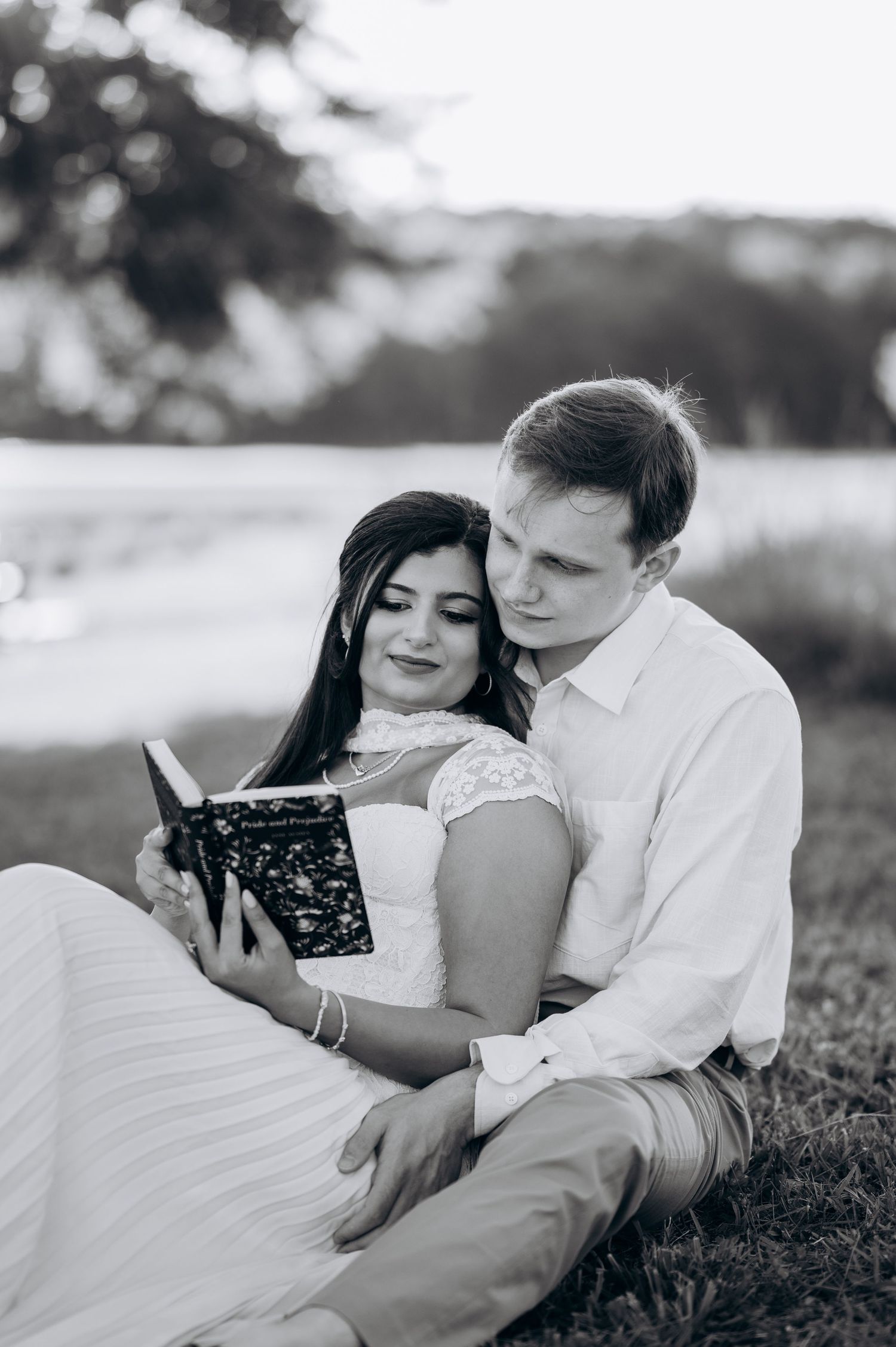 Black and white photo of a couple sitting on grass, woman in white dress reading a book while man embraces her.