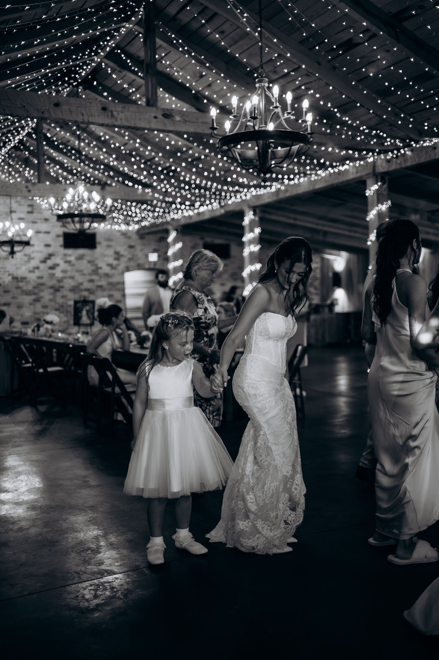 Bride in lace gown dancing with flower girl under string lights at reception.
