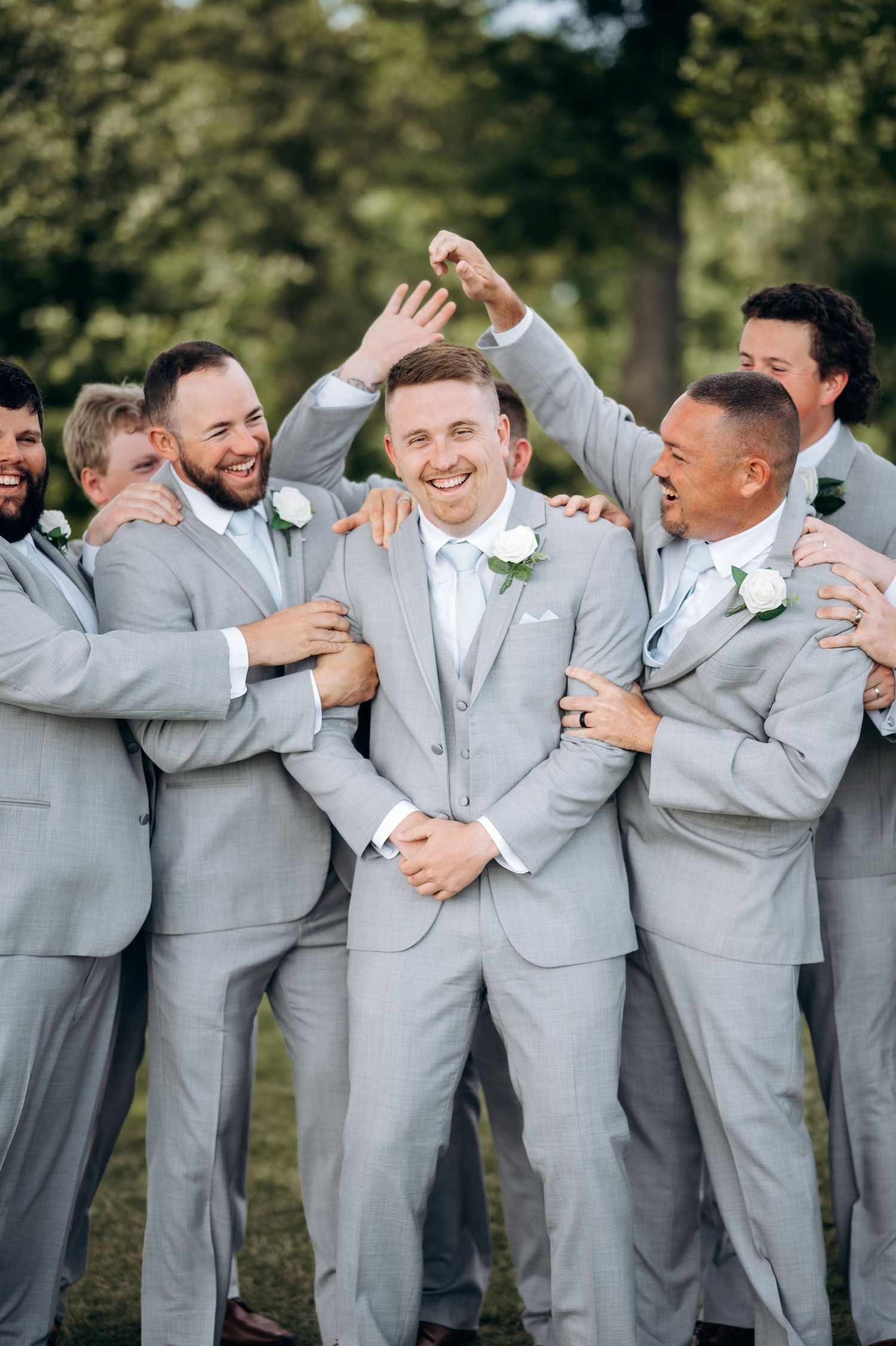 Groom laughs joyfully as groomsmen in matching gray suits playfully surround him during wedding party photos.