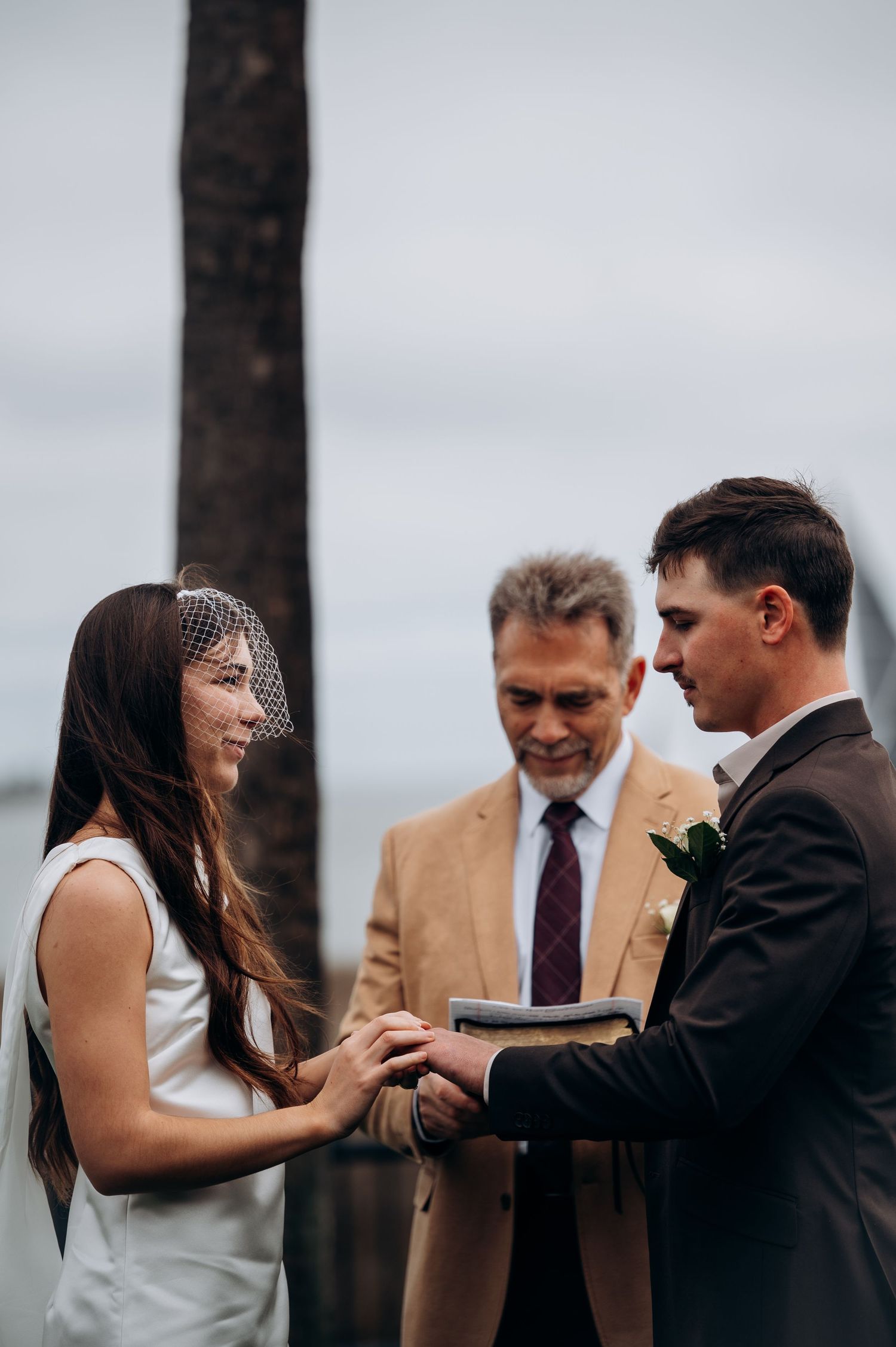 Bride and groom exchange rings with officiant at outdoor coastal wedding ceremony.