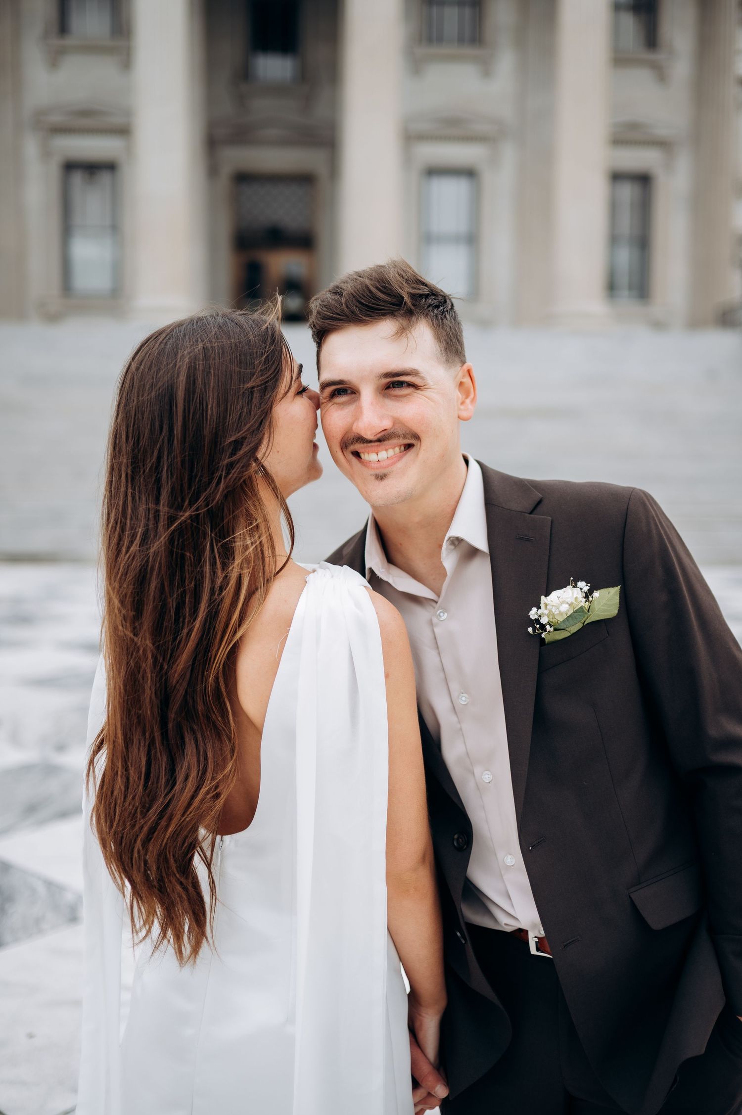 Smiling groom in dark suit with boutonniere receives a kiss on the cheek from bride in white dress outside.