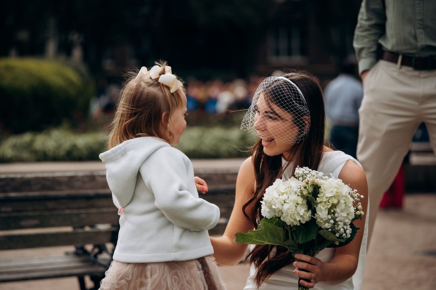 Bride in veil holding white bouquet bends down to speak with flower girl in white jacket at outdoor wedding.
