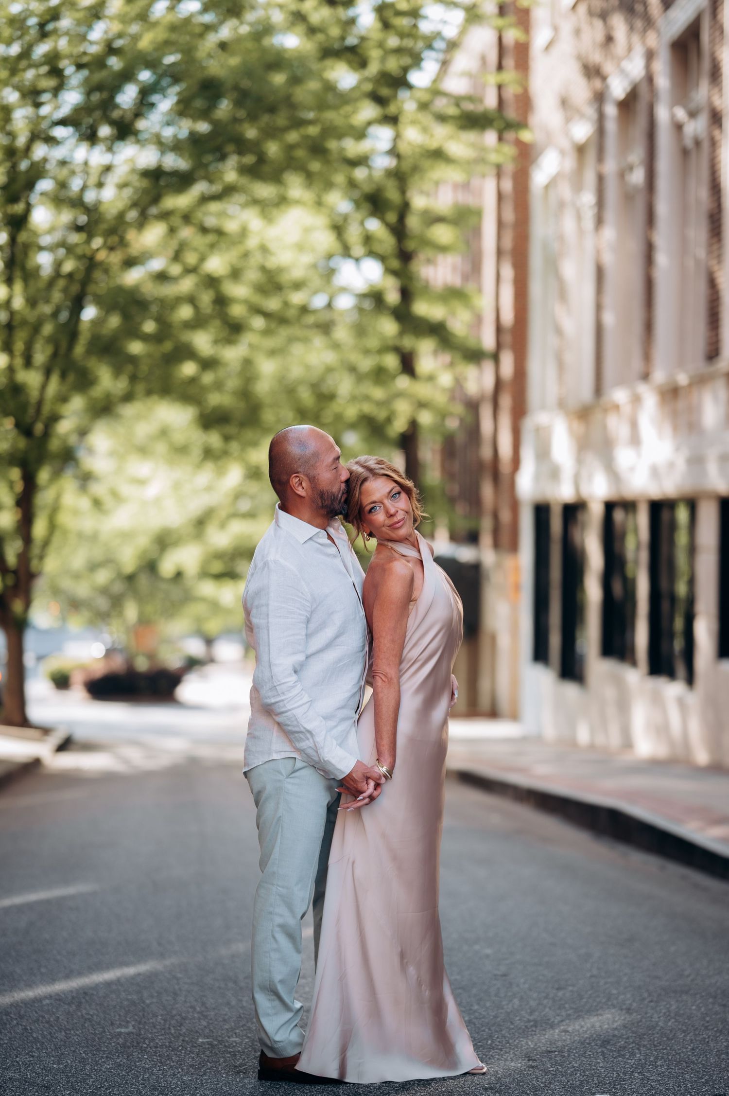 Couple embracing tenderly on a tree-lined urban street, woman in a blush satin gown.