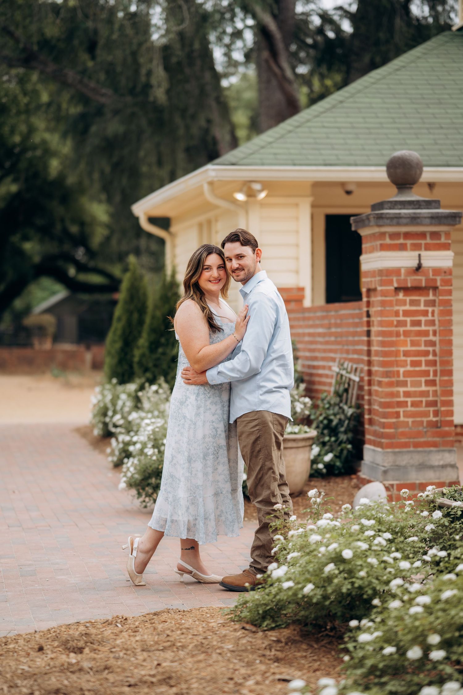 Couple embracing and smiling on a brick path beside white flowers near a charming brick building.