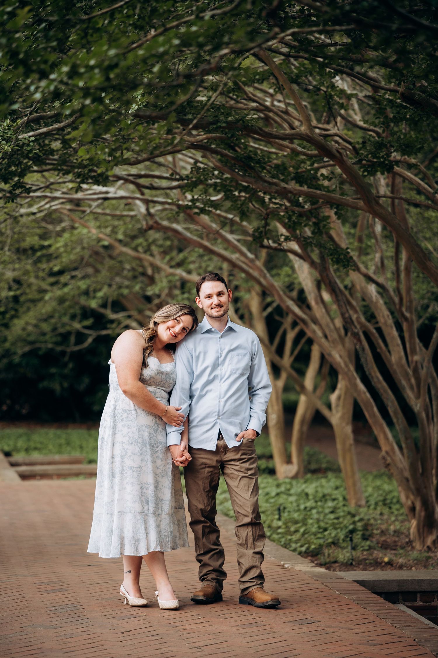 Smiling engaged couple holding hands and posing under arching trees on a brick walkway.