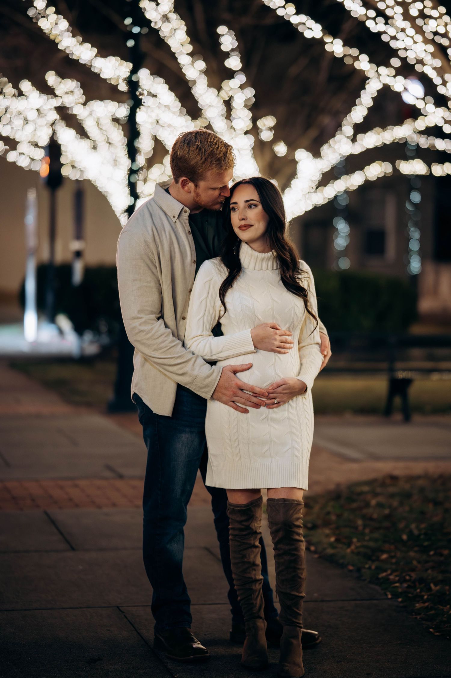 Couple embracing outdoors at night with glowing white lights on trees, woman in white sweater dress showing baby bump.