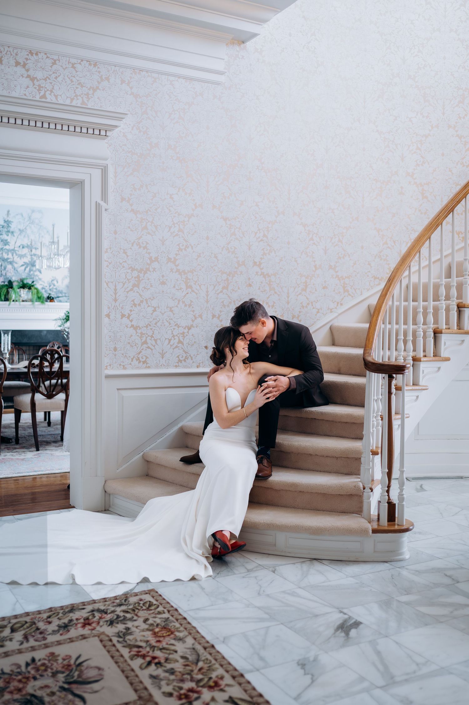 Bride in white mermaid gown and groom in black suit share a kiss on a sweeping marble staircase.