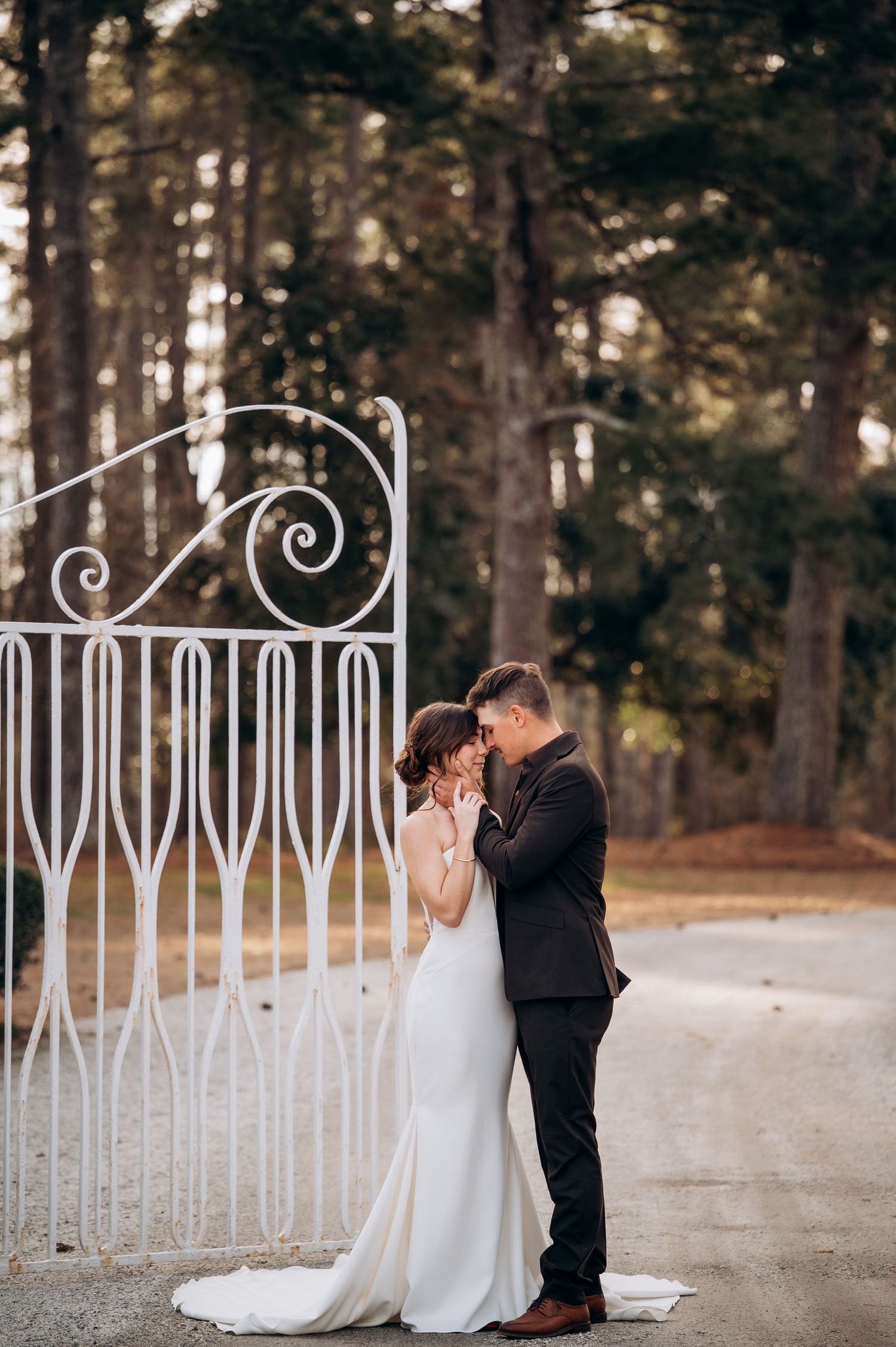 Bride and groom share a romantic embrace before ornate white iron gates surrounded by tall pine trees.