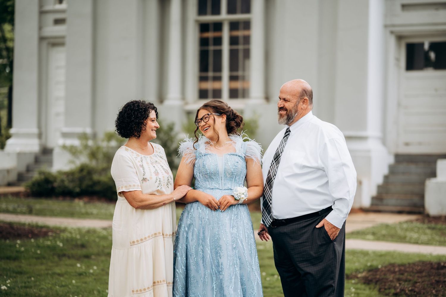 Family of three smiling and laughing together outdoors in formal attire near a white building.
