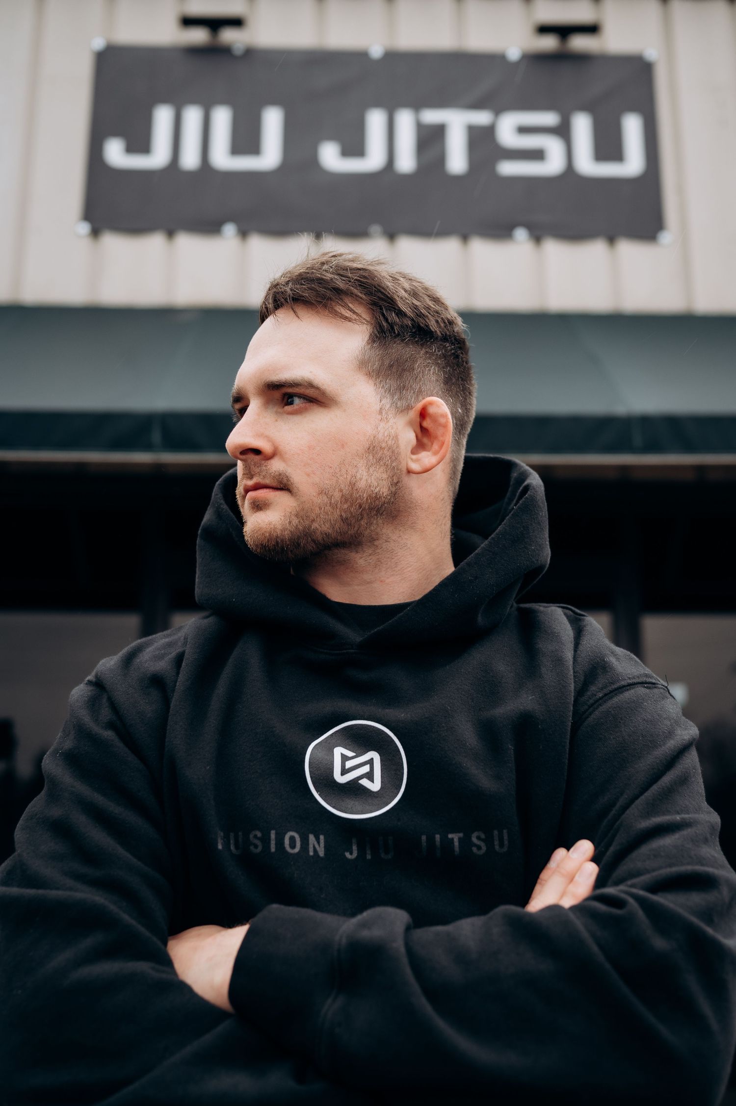 Athletic figure in black hoodie poses against white wall with jiu jitsu branding visible.