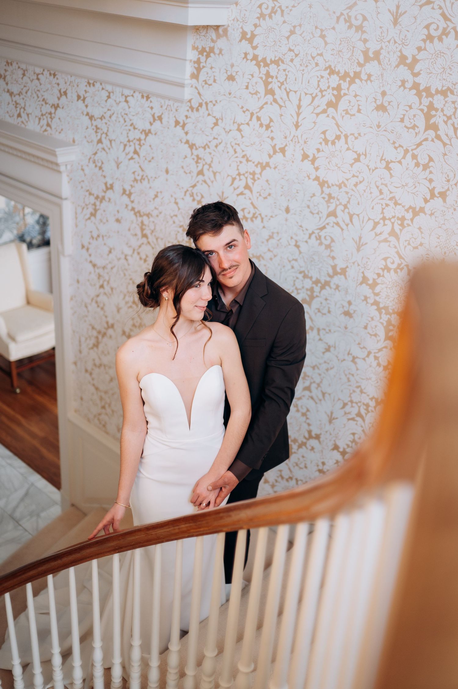 Newlyweds share an intimate moment on a staircase with floral wallpaper in the background.