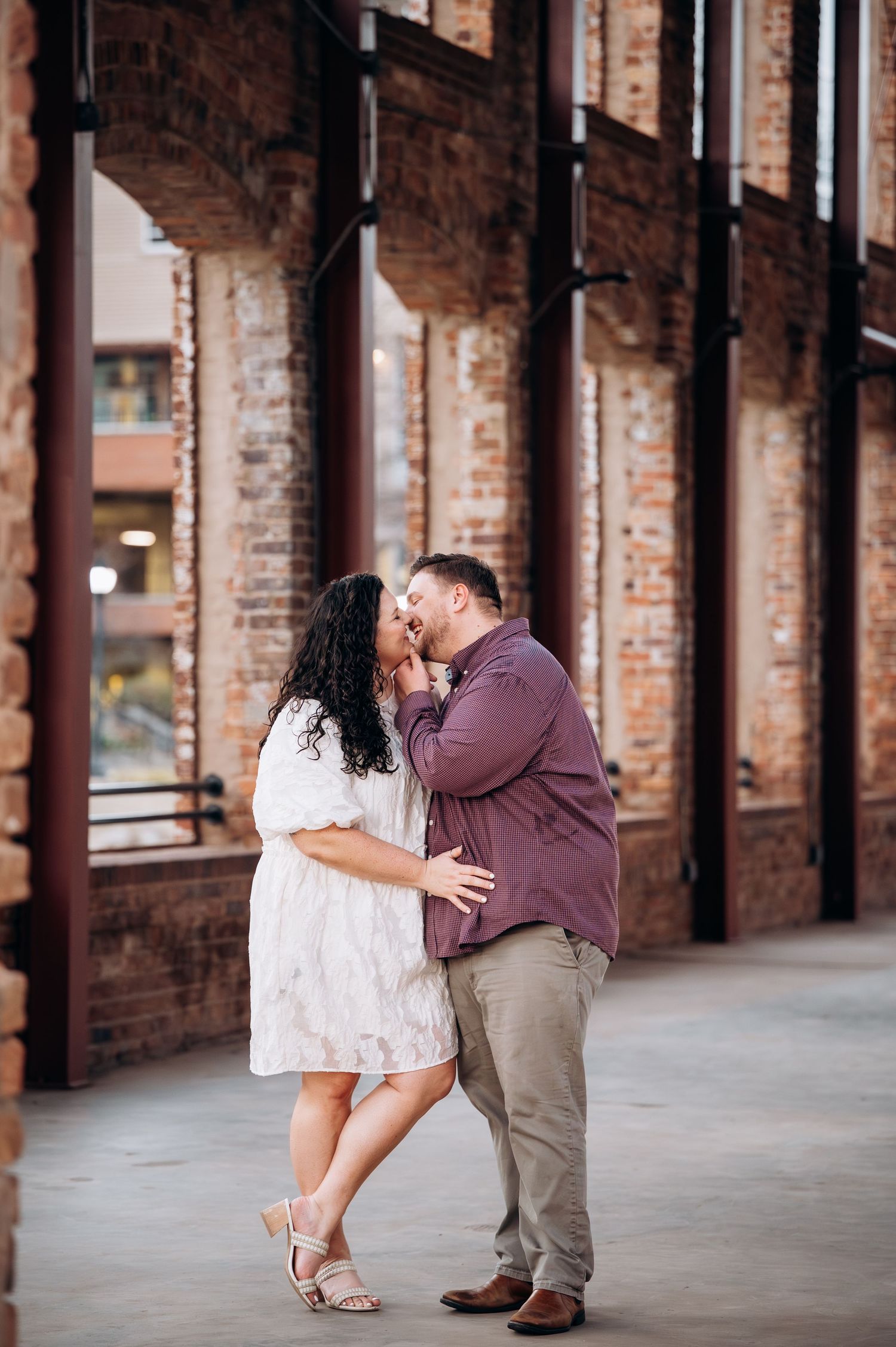 A couple shares an intimate moment while standing in a brick-lined covered walkway with repeating archways.