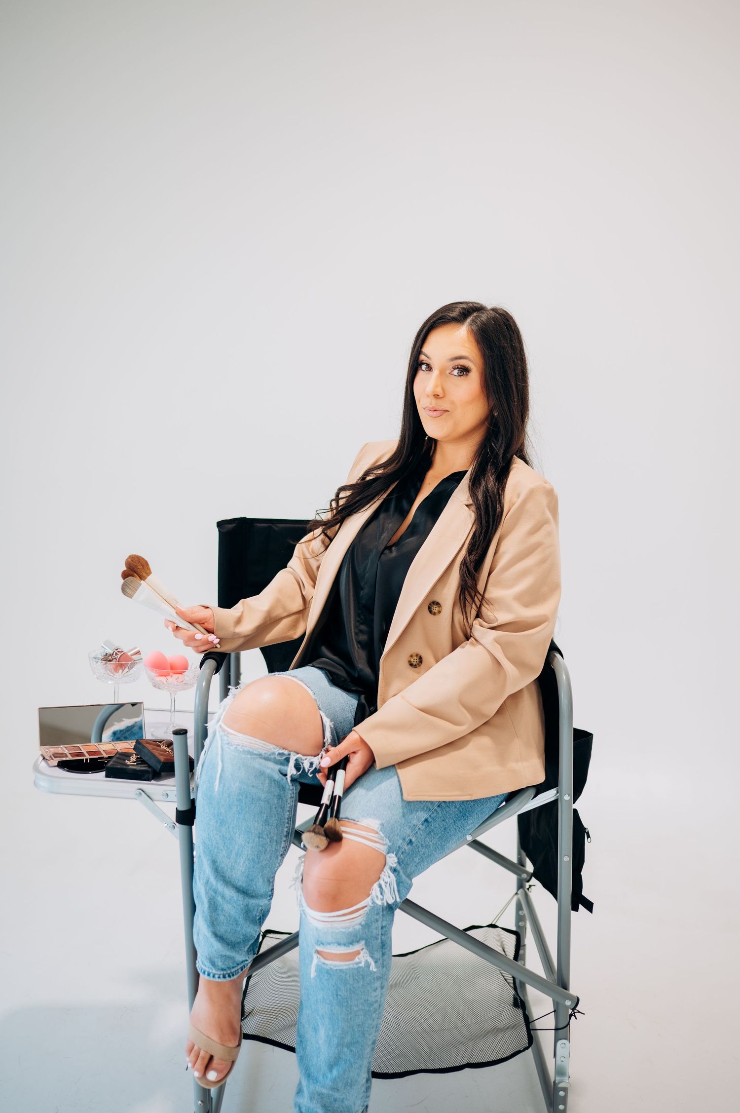A person in a beige blazer and ripped jeans poses in a director's chair against a white backdrop in a photo series.