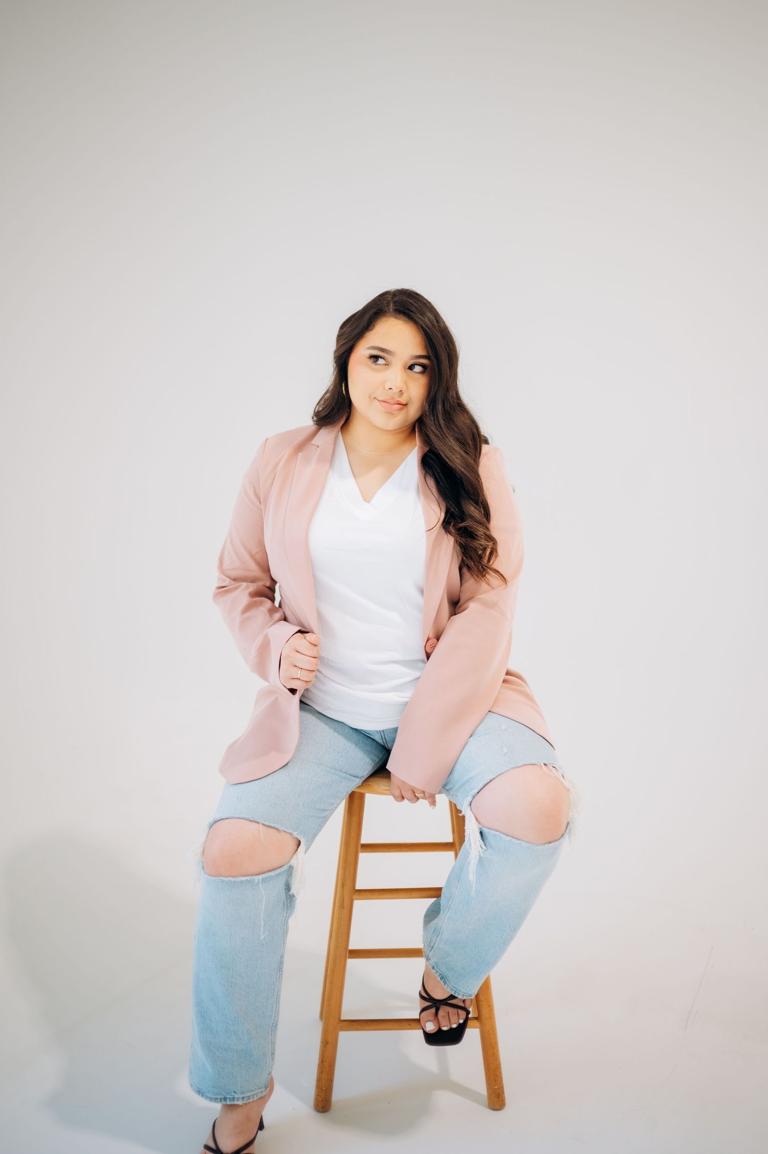 A person wearing a pink blazer and light blue jeans poses casually on a wooden stool against a white background.