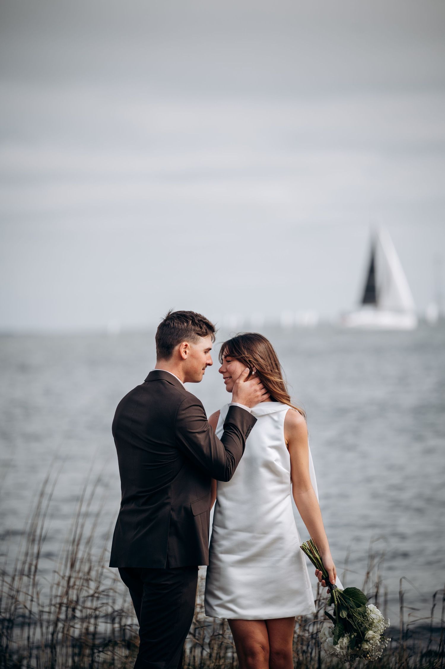 Couple shares tender moments by the waterfront with tall grass and cloudy skies.