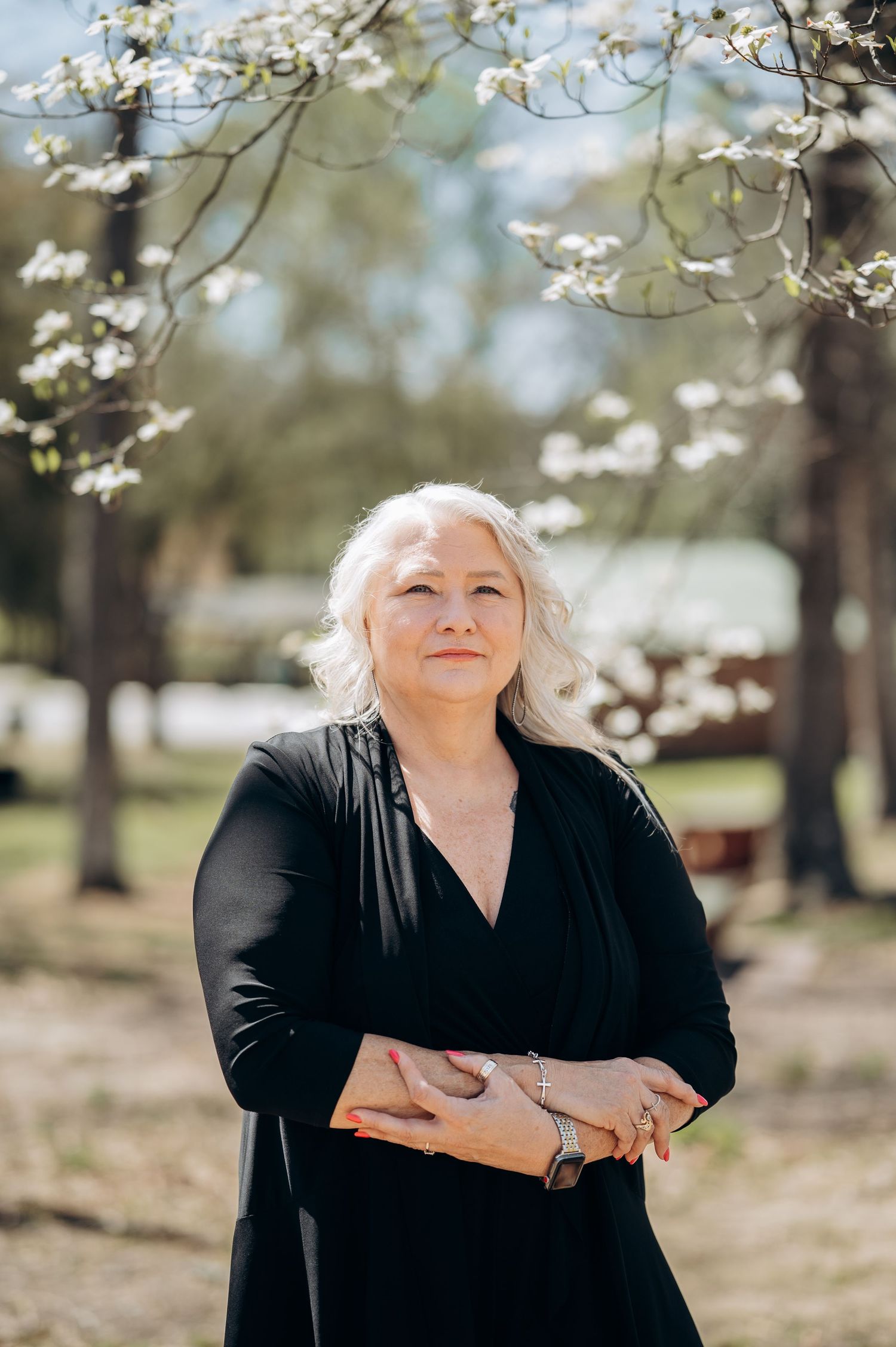 A woman with silver hair in a black dress stands outdoors with arms crossed, blooming trees in the background.