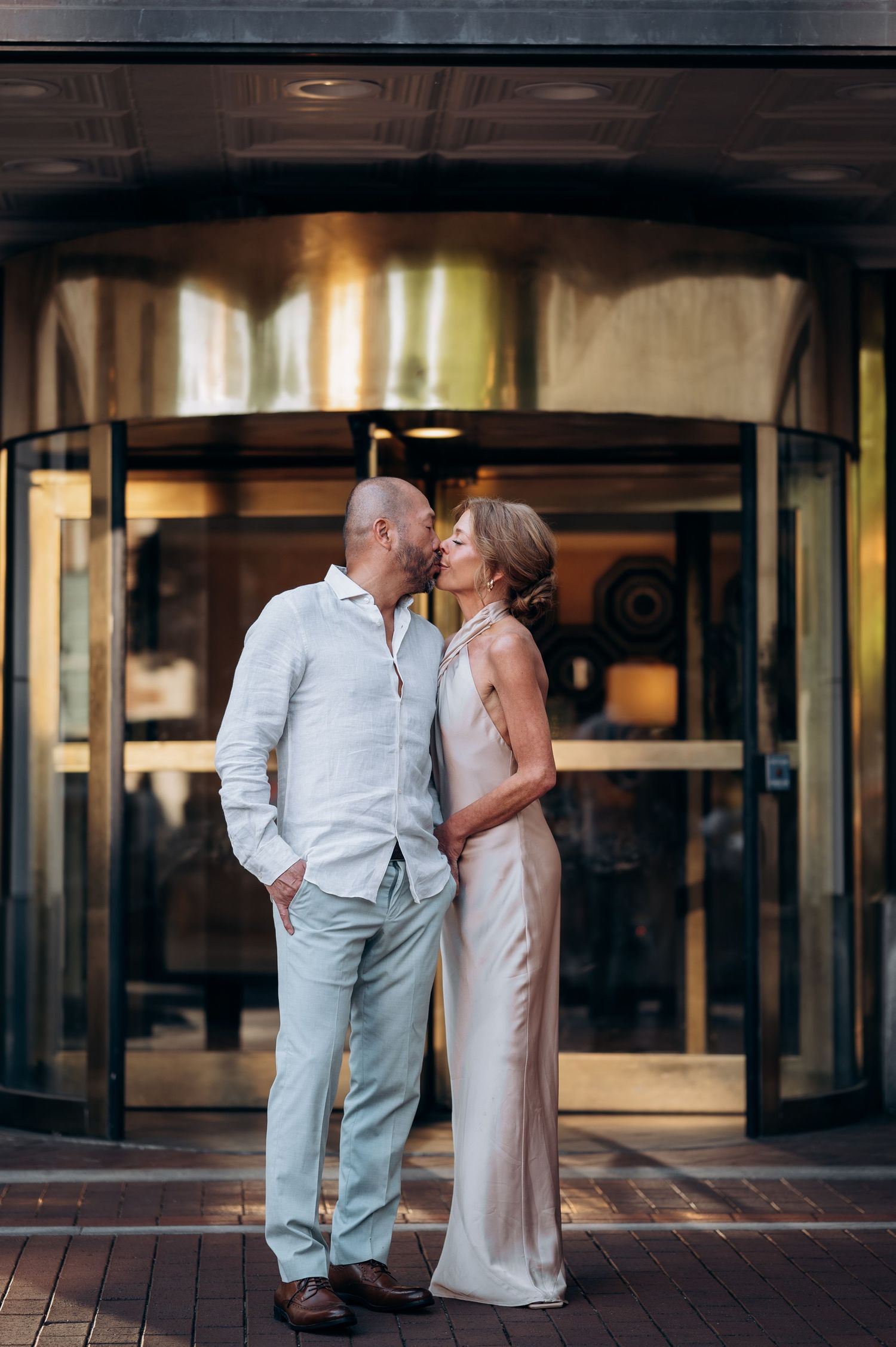 Elegant couple sharing a kiss in front of a revolving door, man in white linen shirt and woman in champagne slip dress.