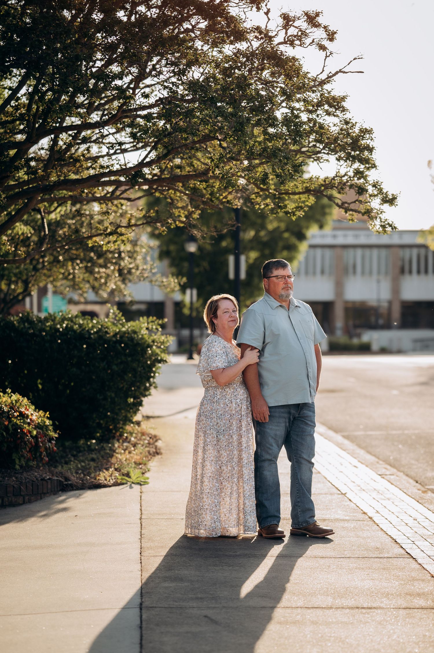Couple standing together on a sunny sidewalk, woman in floral dress and man in blue shirt, surrounded by lush green trees.