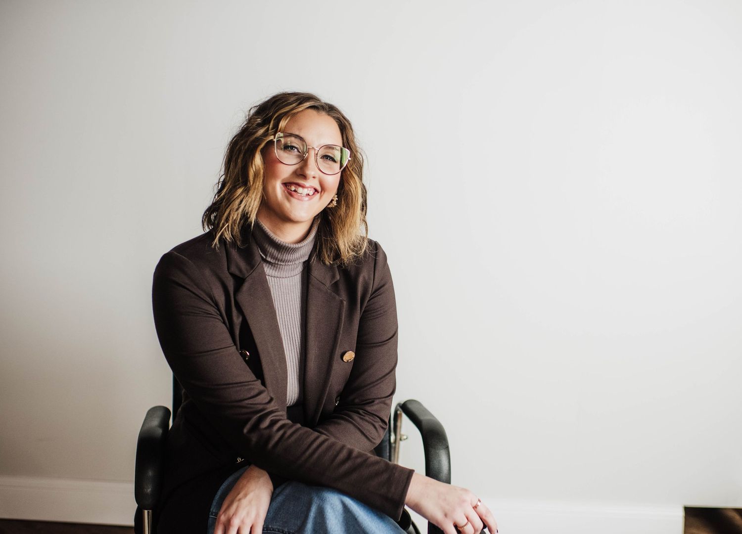 Smiling woman with glasses and wavy hair wearing a brown blazer, seated casually in a chair against a white wall.