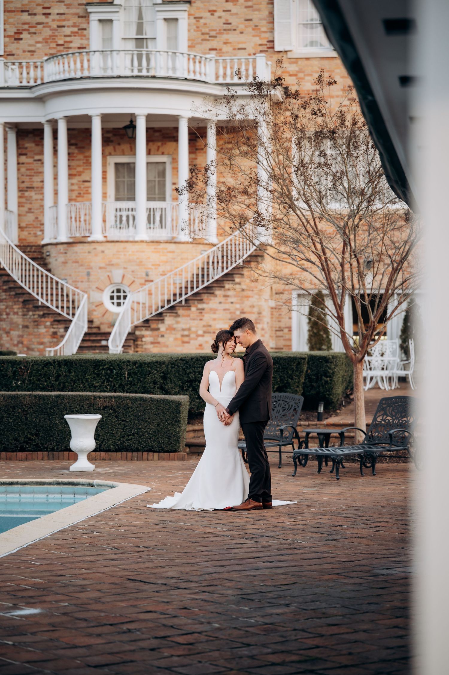 Bride and groom embrace near a pool outside an elegant brick mansion with white balconies on their wedding day.