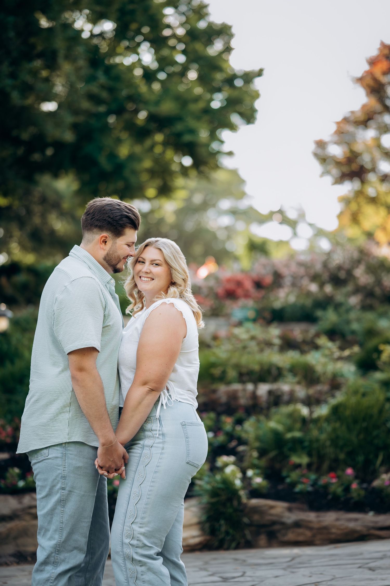 A couple holds hands and embraces in a lush garden, the woman smiling at the camera while the man whispers in her ear.