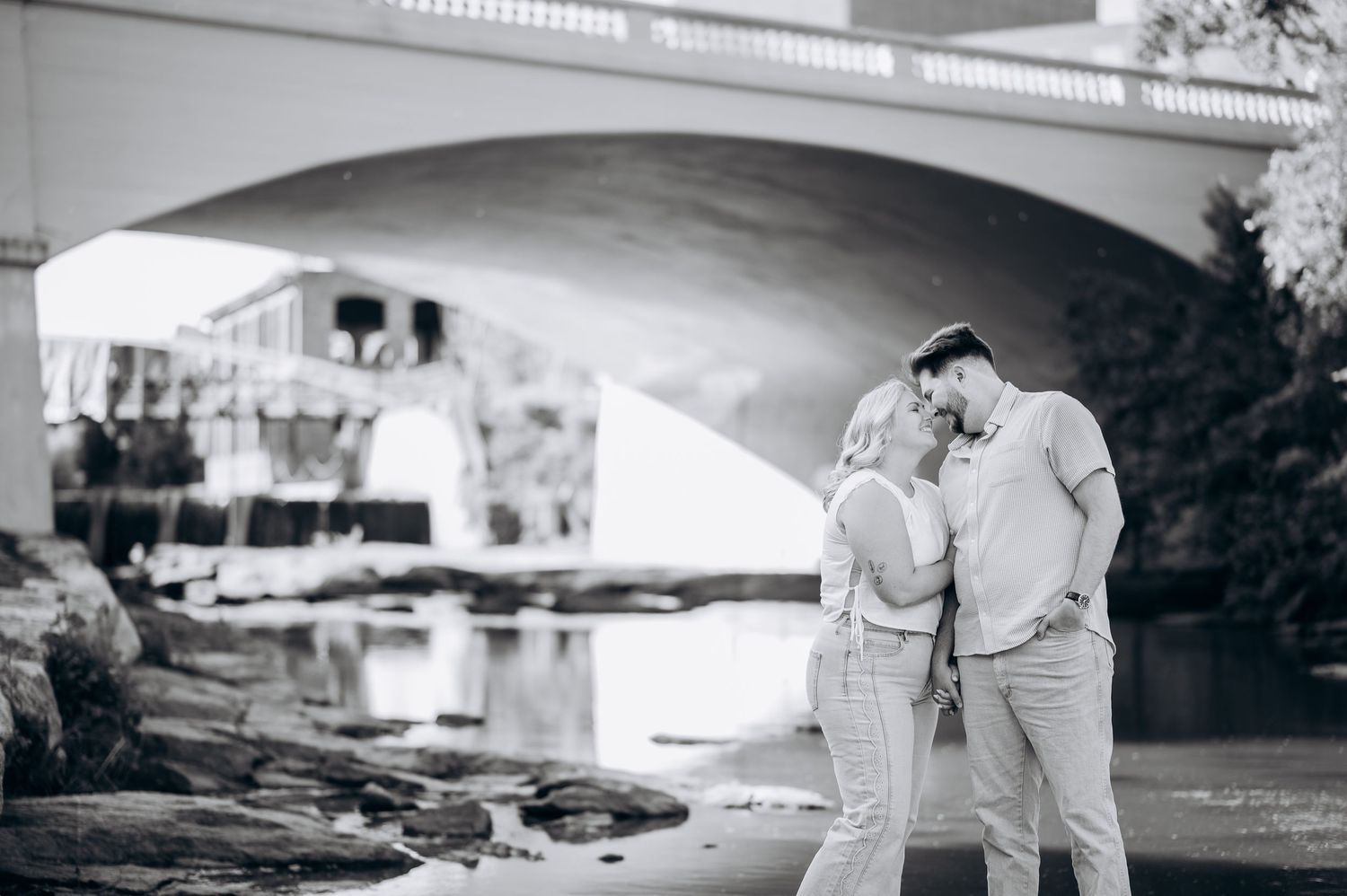 Black and white photo of a couple sharing a romantic moment by a river under a stone bridge.