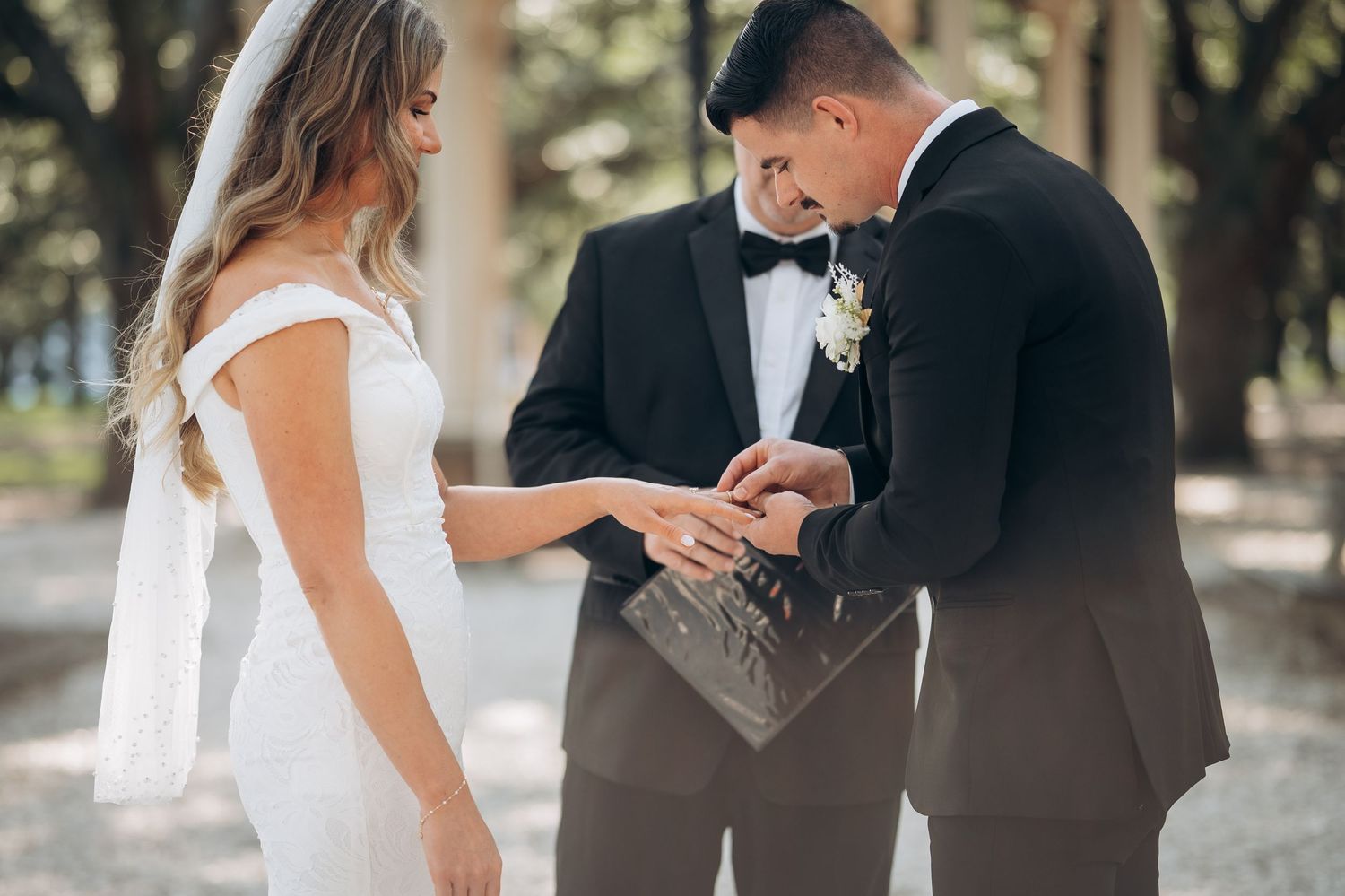Groom placing wedding ring on bride's finger during outdoor ceremony, officiant holding ring box in background.
