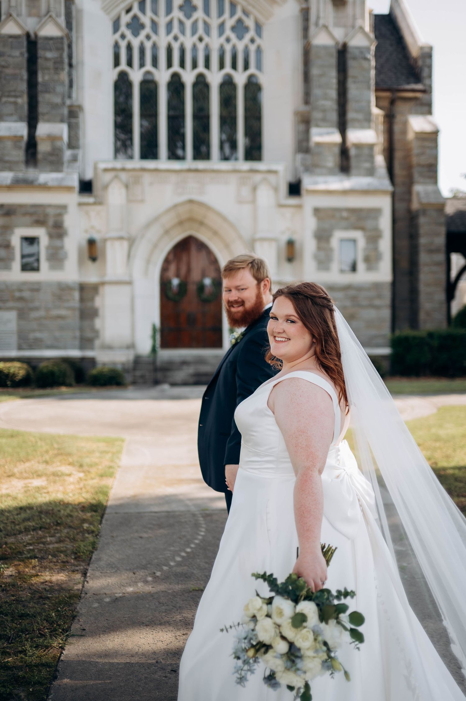 A couple in wedding attire stands in front of a gothic church with stone architecture.