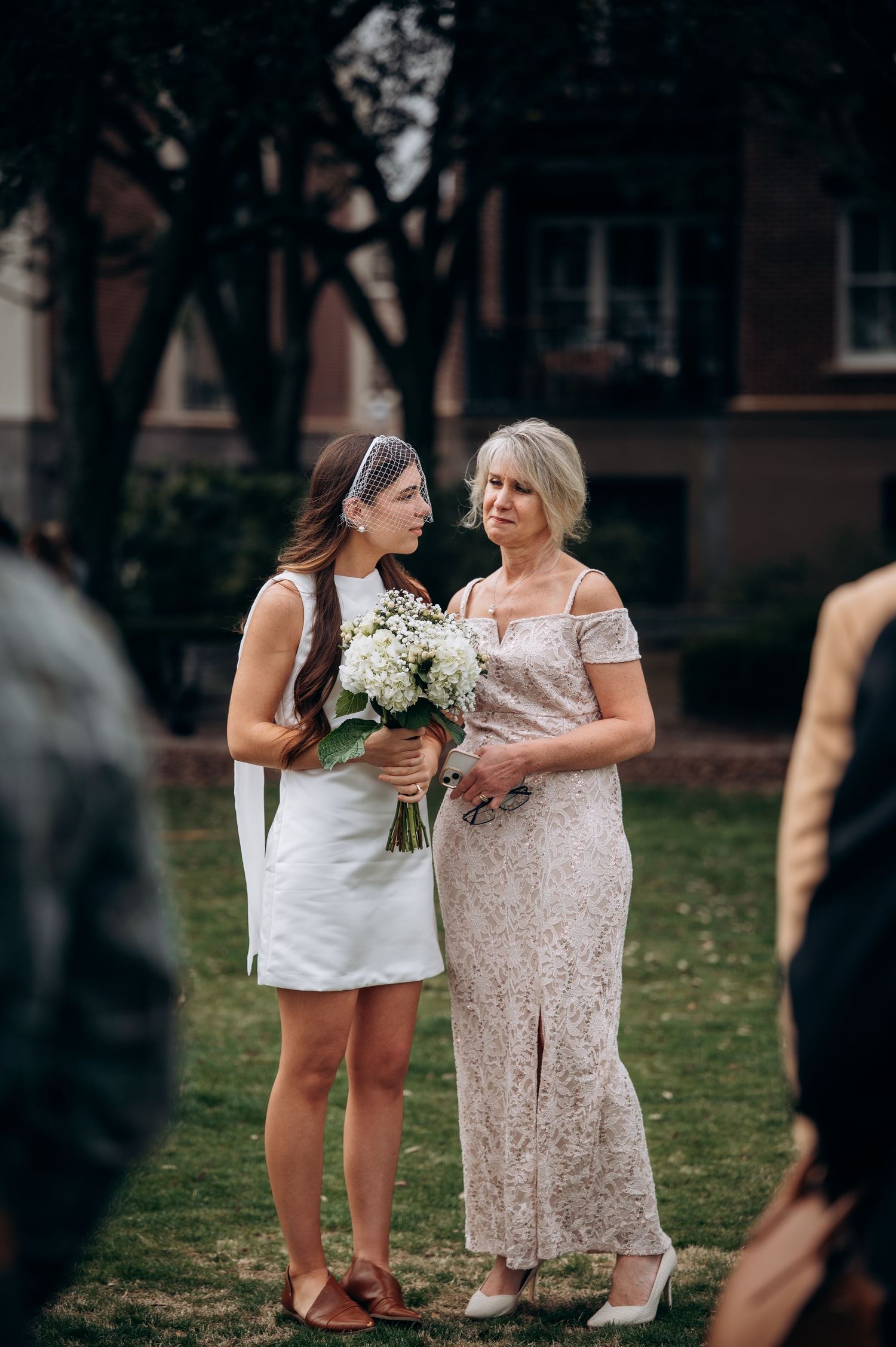 Bride in short white dress holds bouquet beside older woman in lace gown at outdoor wedding.