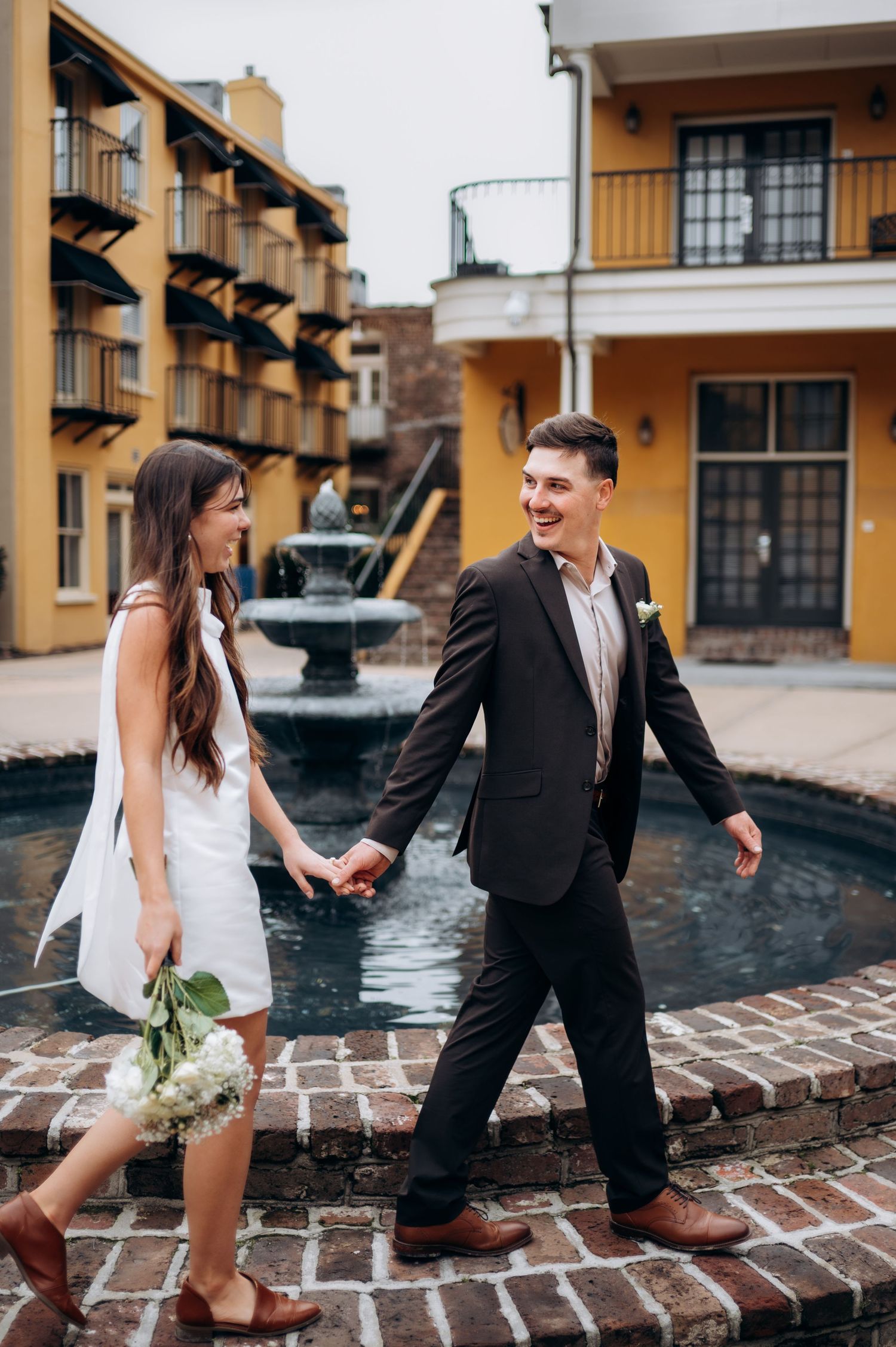 Happy newlywed couple holding hands and laughing near a courtyard fountain, bride holding white flowers.