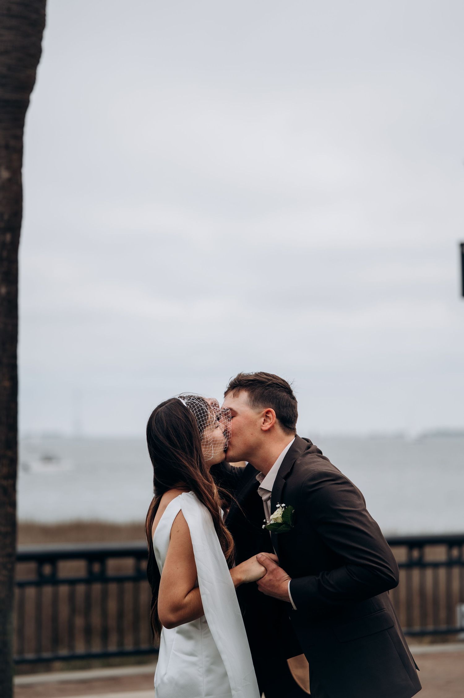 Bride and groom sharing first kiss by the waterfront on their wedding day under cloudy skies.