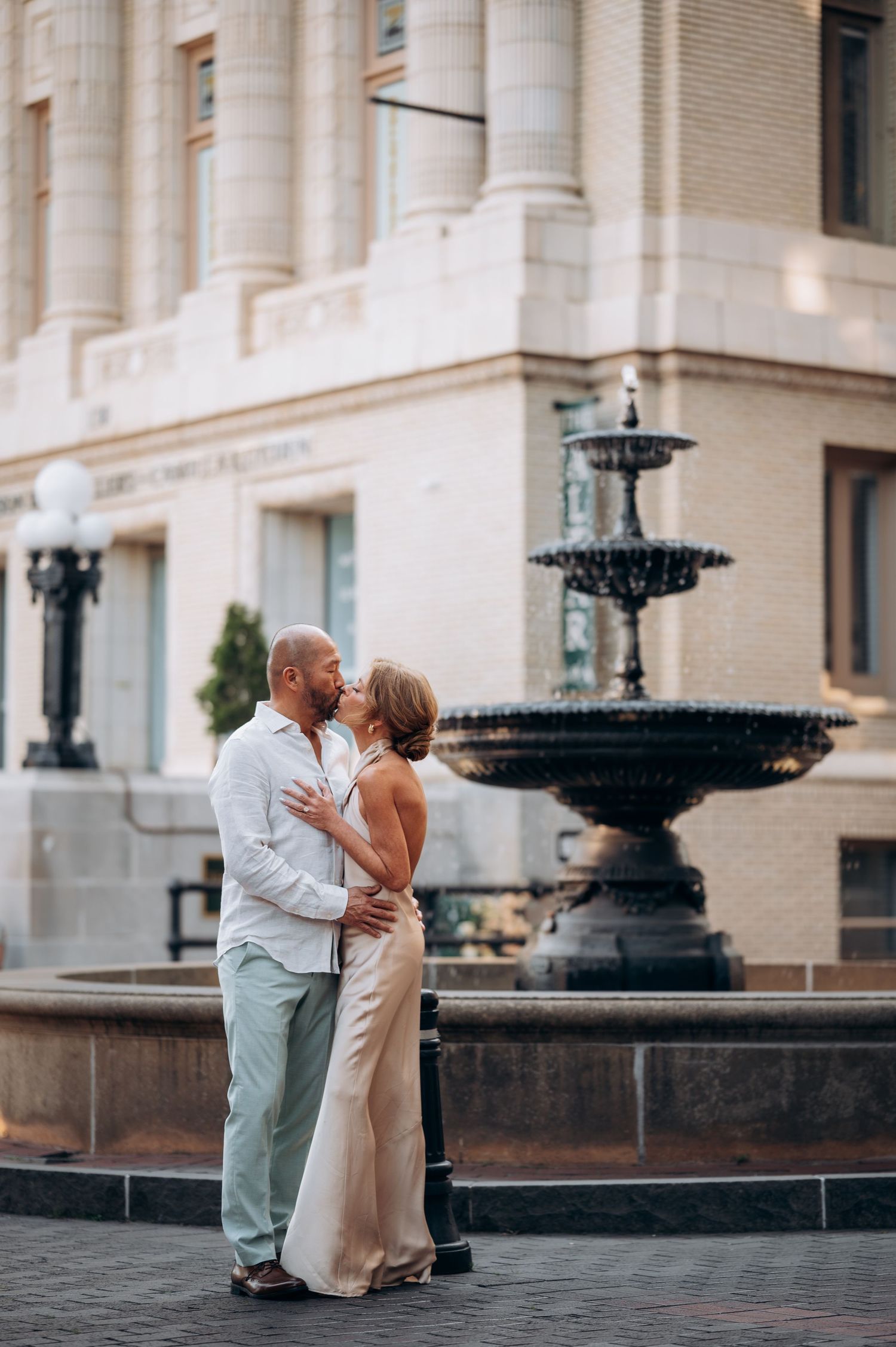 Wedding couple sharing a kiss beside a classic stone fountain in a city plaza.