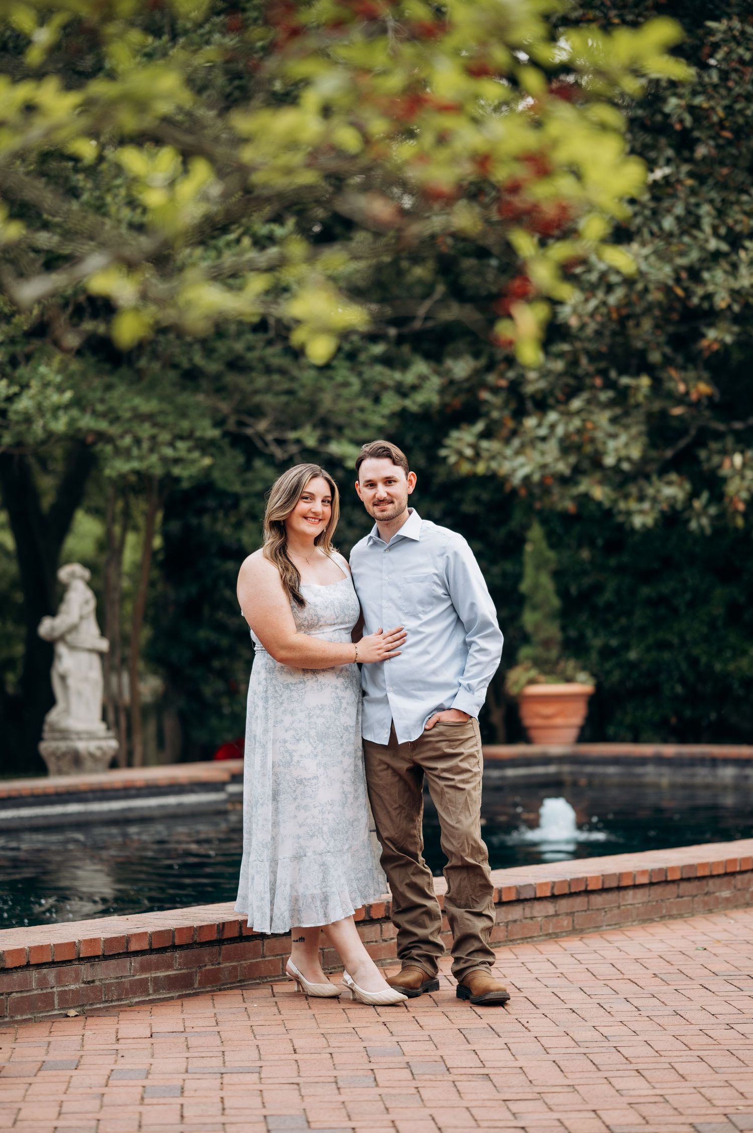 Smiling couple posing together on a brick patio beside a garden fountain surrounded by lush trees.