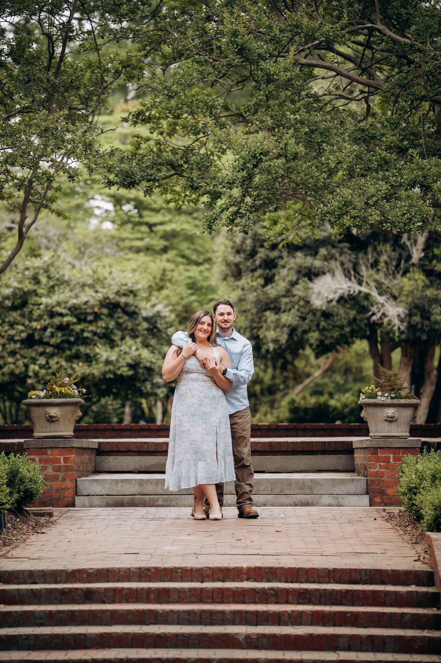 Happy couple posing on brick steps surrounded by trees and decorative urns in a garden.