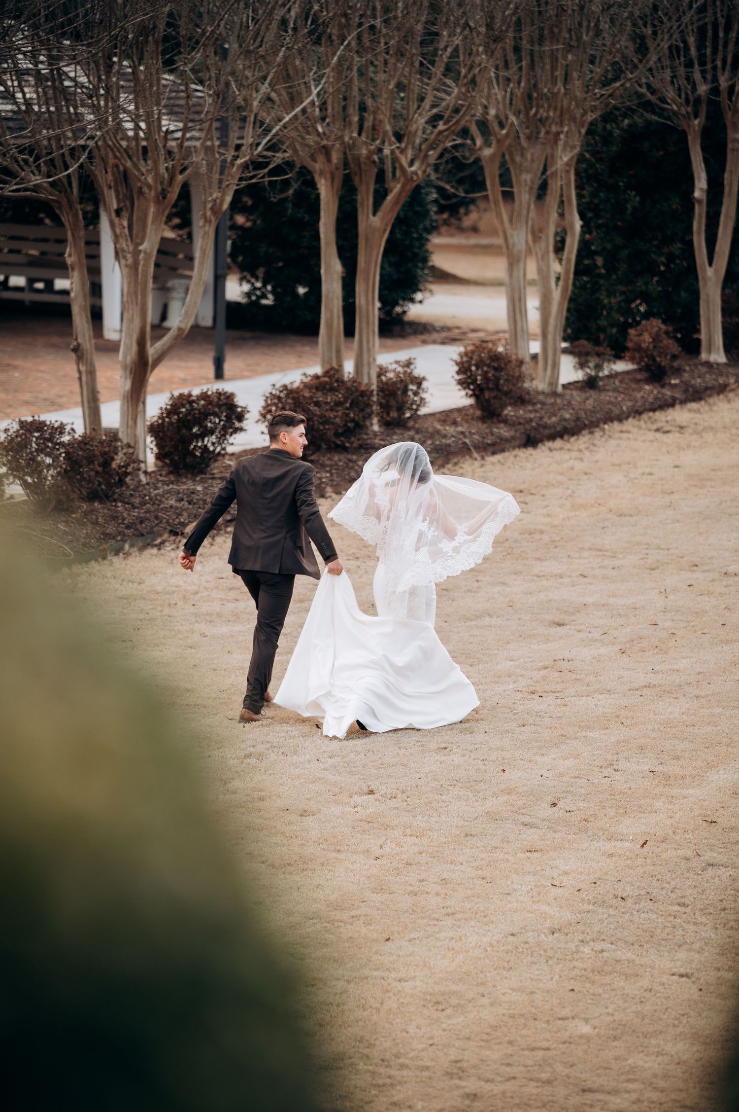 Groom carrying bride's flowing veil and train as couple walks through tree-lined wedding venue grounds.